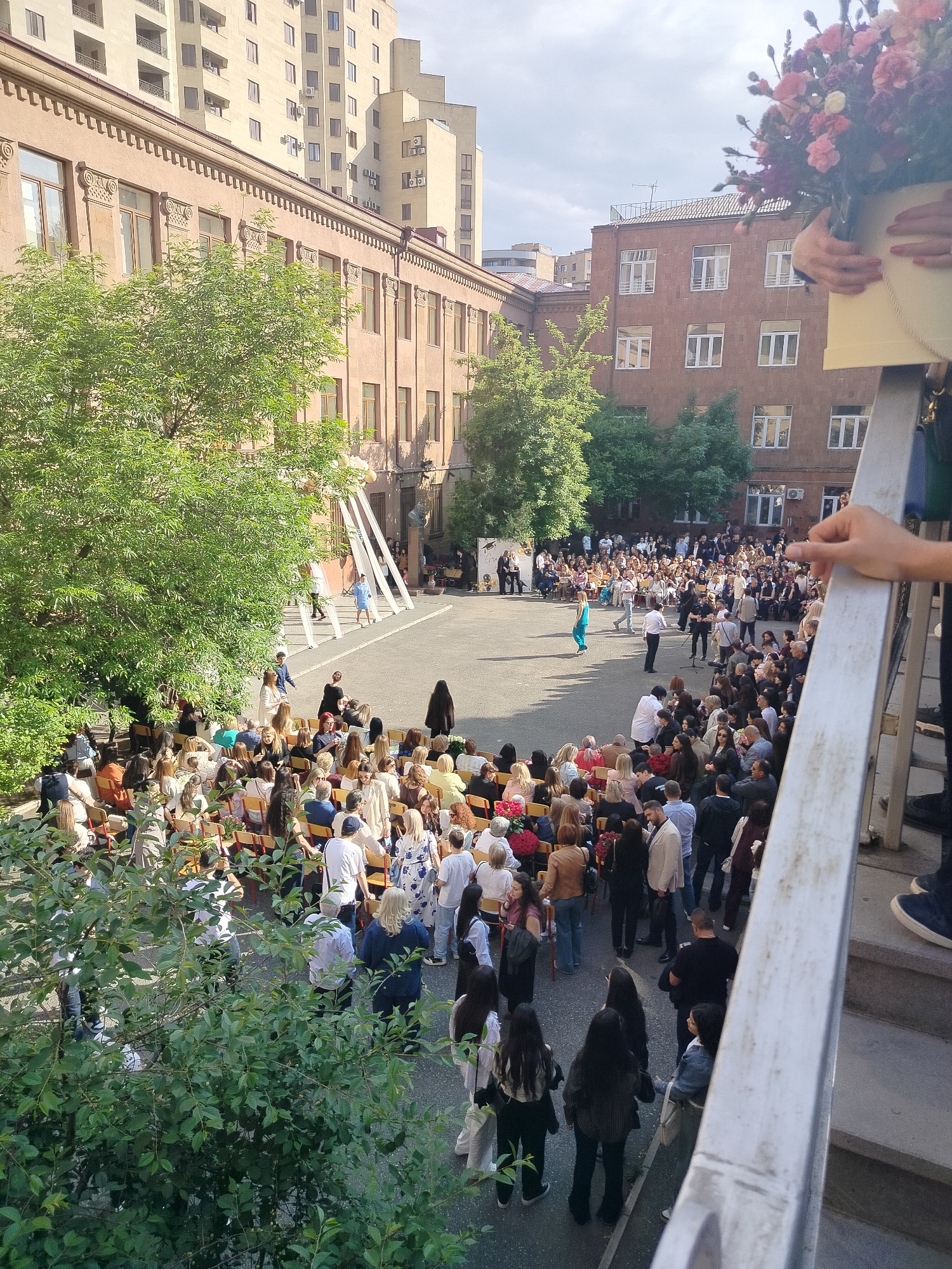 A high-angle view shows a large gathering of people in the courtyard of a building, likely a graduation ceremony or similar event. The courtyard is enclosed by several multi-story buildings of similar architectural style, suggesting a school or institutional complex. The image was most likely taken in Yerevan, Armenia.
Rows of chairs are arranged on the left side of the courtyard, occupied by attendees. A group of people are standing in the middle, with some facing a makeshift stage or platform adorned with white fabric. The crowd is composed of individuals of various ages and appearances, with most of them appearing to be adults.
On the right side, a person is partially visible on a staircase, holding a bouquet of flowers in a decorative container. The perspective suggests the photograph was taken from an elevated position, potentially from the staircase itself. The environment indicates an urban setting, with trees and greenery interspersed among the buildings. The overall impression is of a formal or ceremonial gathering in an educational or institutional environment.