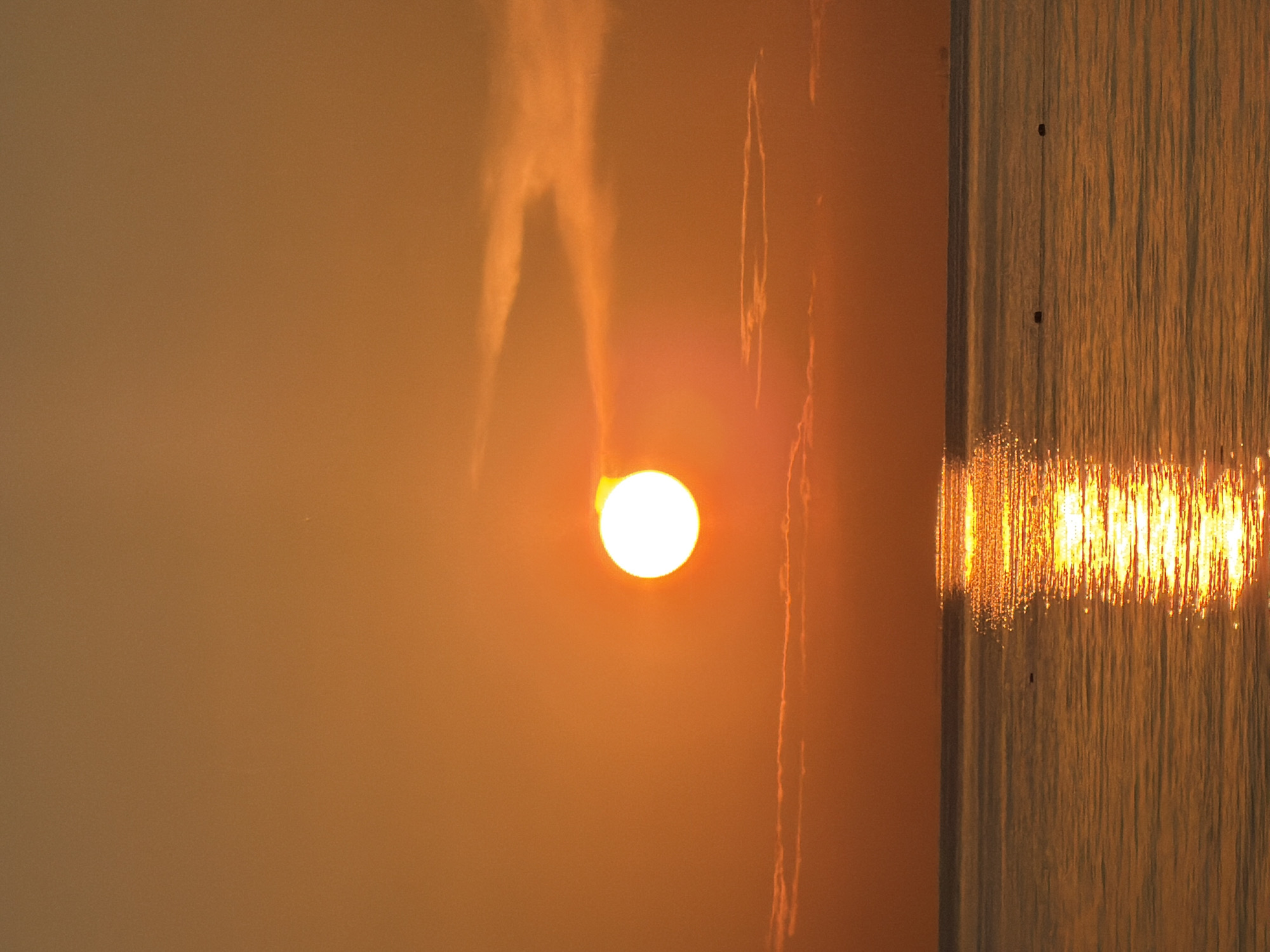The image shows a vibrant sunset over the Black Sea, with the sun appearing as a bright, glowing orb low on the horizon. The sky is a warm gradient of orange and yellow, with a few wispy clouds illuminated by the setting sun. The calm waters of the sea reflect the golden hues of the sky, creating a shimmering path of light that stretches towards the viewer. A thin line of clouds or land can be seen in the distance, dividing the sky and sea. There are at least two small, dark objects floating on the water's surface, which appear to be buoys. The scene evokes a sense of tranquility and natural beauty. The location is the city of Sochi, Russia.