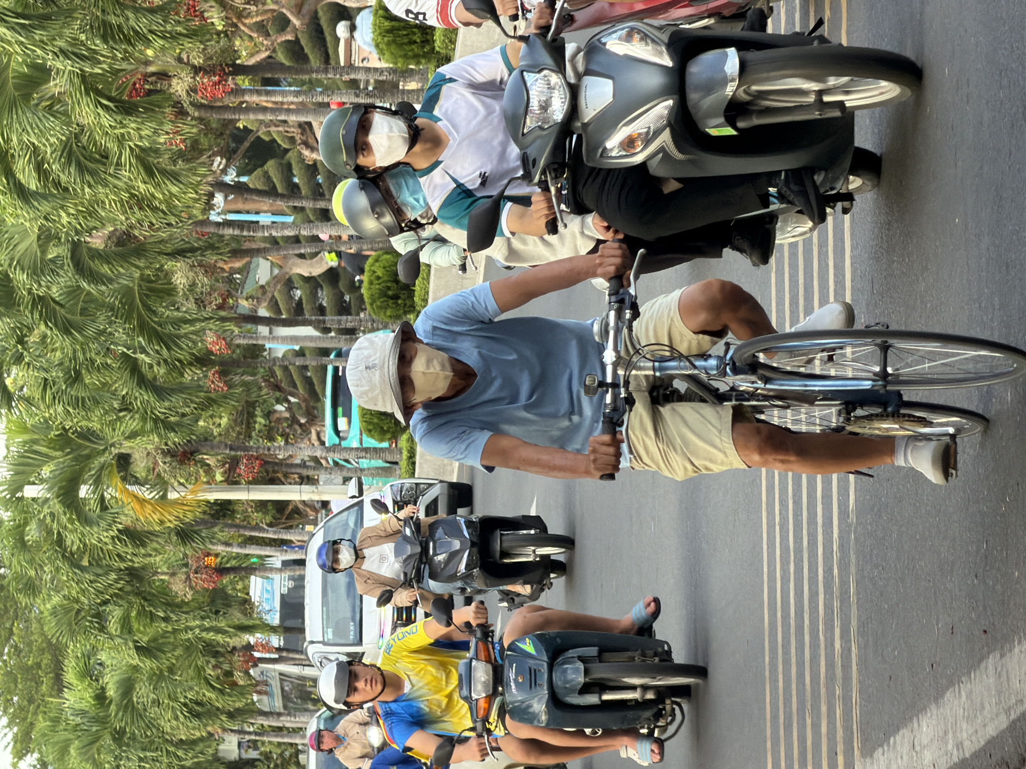 The image depicts a street scene in Nha Trang, Vietnam, likely during the daytime given the lighting. The foreground features two individuals on bicycles and several people on motorcycles, navigating a multi-lane road lined with palm trees. All visible individuals are wearing face masks.
On the left, a young man wearing a yellow and blue t-shirt and light-colored shorts rides a dark green motorcycle. He is wearing a white helmet and a light blue face mask. Next to him, partially obscured, is another person on a motorcycle.
In the center foreground, a man wearing a light blue t-shirt, khaki shorts, and a light-colored hat rides a bicycle. He is wearing a beige face mask. To his right, a person on a dark gray motorcycle is visible from the chest down. This individual wears dark pants and black shoes, and a portion of their white and green patterned shirt is visible. A person behind them on the same motorcycle wears a light gray hoodie and a light blue face mask.
Behind these individuals, a white car with "TAXI" written on it is stopped in traffic. A person wearing a brown jacket and a blue face mask is riding a dark gray motorcycle in front of the taxi. Further back, other vehicles and people on motorcycles are present, along with more palm trees and what appear to be manicured bushes, suggesting a well-maintained urban environment. The road surface is asphalt with yellow lane markings. The overall impression is of a typical traffic scene in a tropical coastal city.