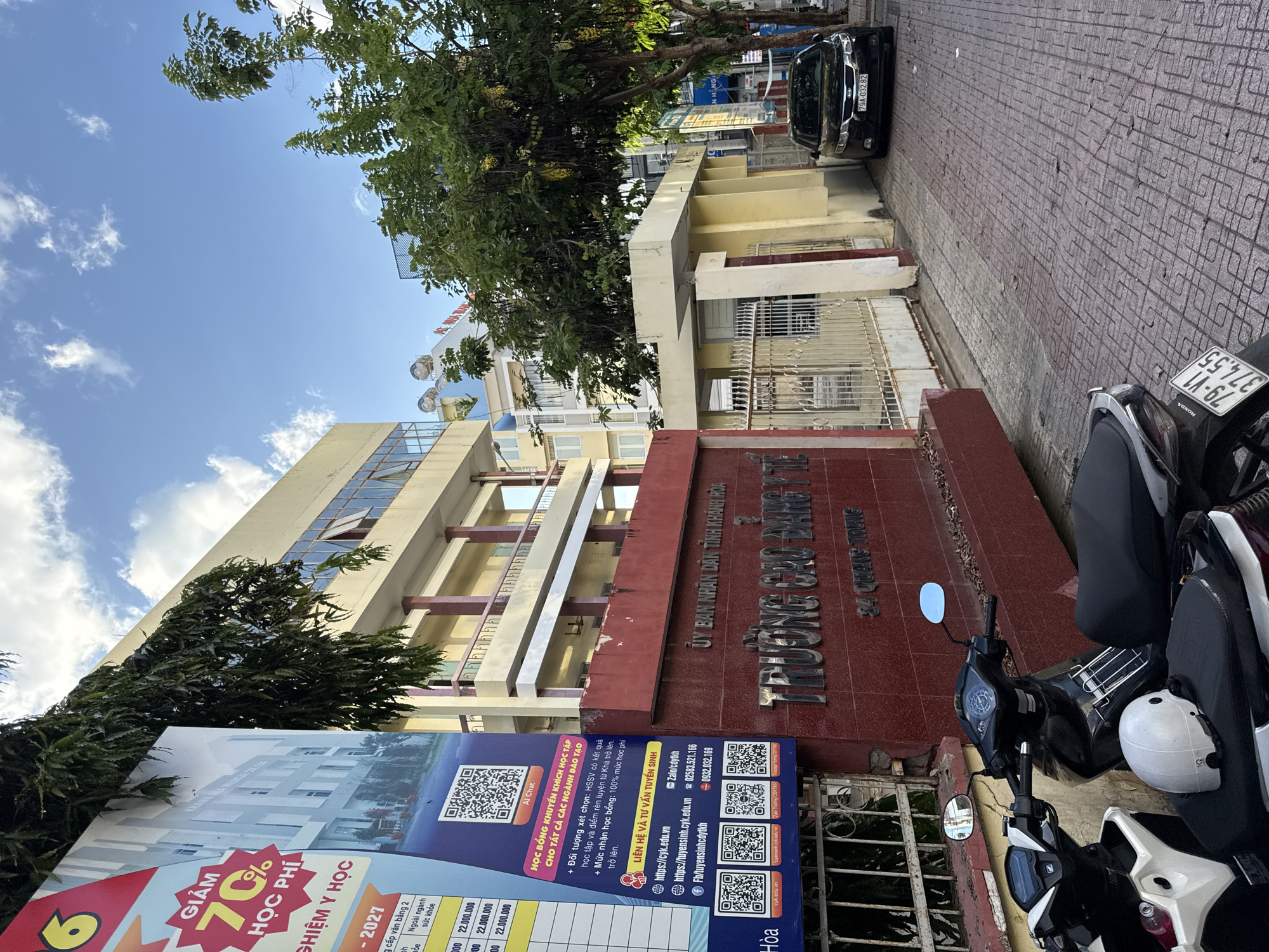 The image shows a street view in front of a building with Vietnamese signage. On the left is a large billboard advertising educational programs, featuring a 70% discount on tuition fees for the academic year 2027. The billboard also displays QR codes for various contact methods and admission information, including websites and phone numbers. The building itself appears to be a school or college, indicated by the prominent red sign that reads "TRƯỜNG CAO ĐẲNG Y TẾ" (Nha Trang Medical College) and the address "34 QUANG TRUNG". To the right of the entrance gate, a black Kia sedan is parked on the street. In the foreground, several motorcycles are parked along the sidewalk. The street is paved with patterned tiles, and trees with green foliage line the walkway. The sky is blue with scattered white clouds. The overall scene depicts an urban environment in Nha Trang, Vietnam, likely during daylight hours given the lighting conditions.