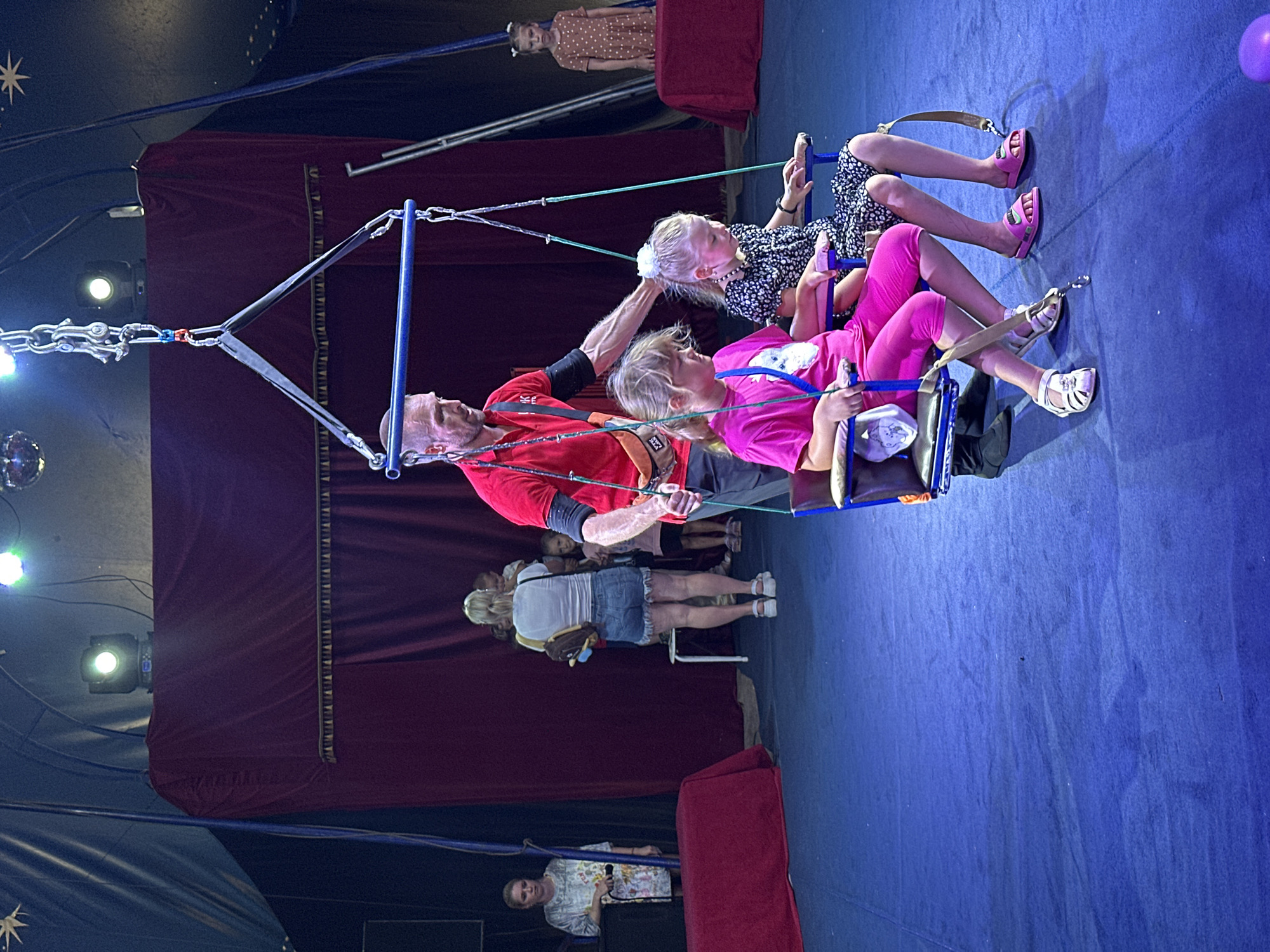 This is a medium shot photograph taken at a circus performance in Novoaltaysk, Russia. The scene is dominated by a blue circus ring floor and a dark red velvet curtain backdrop. A trapeze apparatus hangs from the ceiling, with a man in a red shirt and black sleeves holding onto one of the ropes. Two young girls are seated in a trapeze seat, also suspended by ropes. The girl on the left wears a bright pink t-shirt and pink leggings, and the girl on the right wears a black and white patterned dress. The man appears to be assisting or observing the girls. In the background, several adults and children stand near the curtain, some of whom are watching the performance. The lighting suggests an indoor evening event, with spotlights illuminating the performance area.