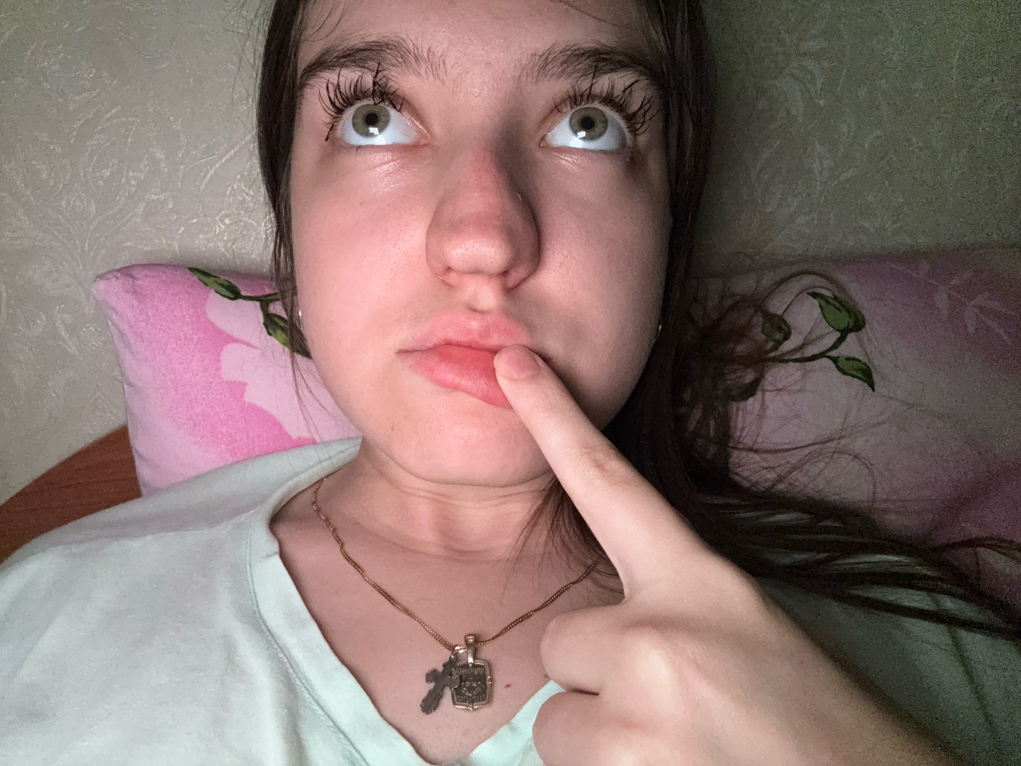 A young person is captured in a close-up, selfie-style shot, their face filling most of the frame. They are looking upwards, with their eyes wide open and pupils dilated, giving them an intense and possibly surprised or inquisitive expression. Their mouth is slightly open, with the index finger of their right hand gently touching their lower lip. The person is wearing a light mint green t-shirt, and a delicate gold necklace with two pendants is visible around their neck. The background consists of a textured wallpaper, and parts of two pillows are visible; one is pink with a floral pattern, and the other appears to be green. The lighting suggests an indoor setting, possibly during the evening or in a dimly lit room. The overall mood is intimate and candid, with a sense of introspection or playful curiosity conveyed by the subject's gaze and gesture. There is no visible text in the image. The setting hints at a personal space, likely a bedroom, in Zavolzhye, Russia.