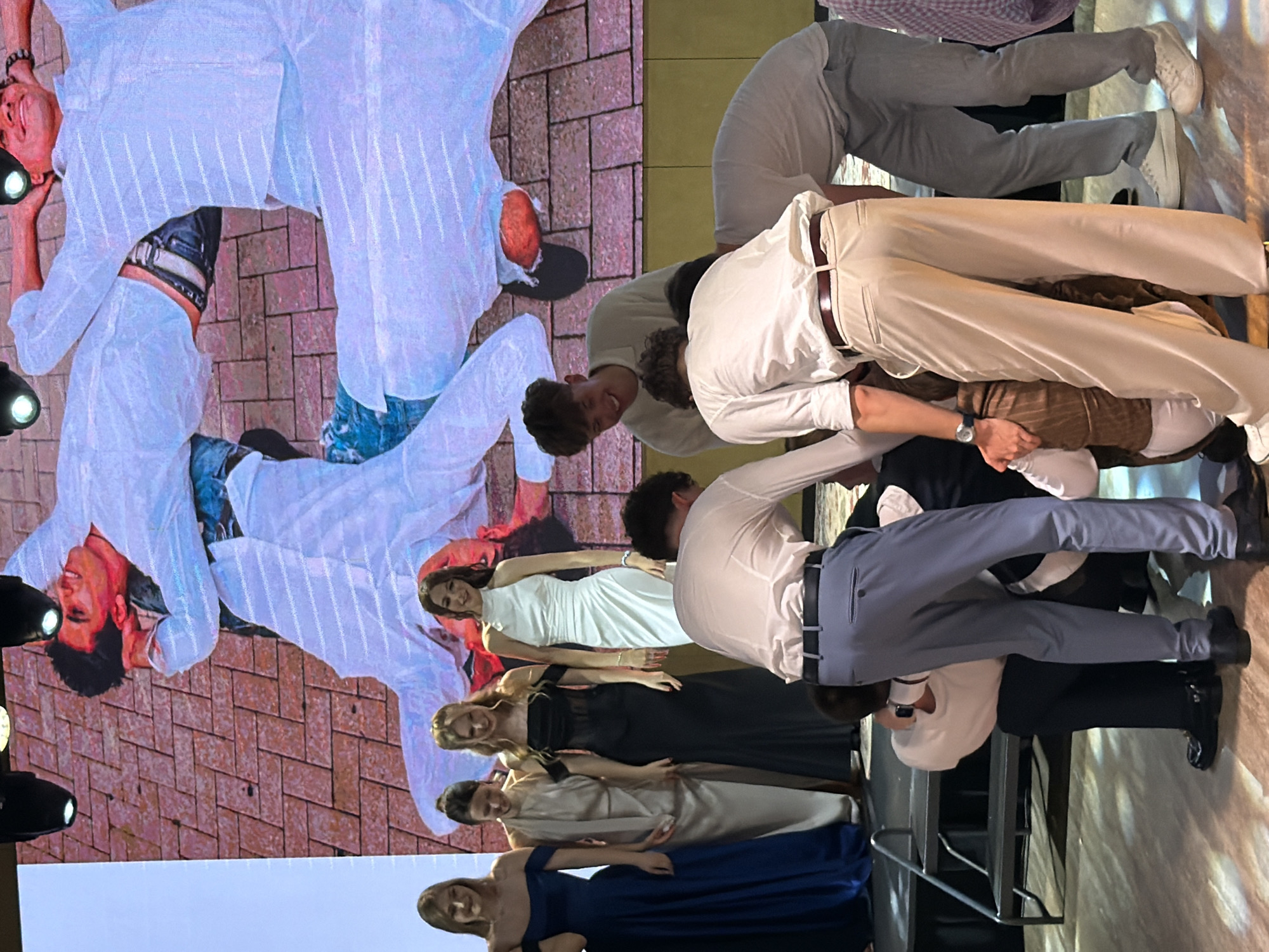 This media file shows a group of people, mostly young adults, in what appears to be a performance or a stylized photoshoot. The subjects are dressed in a mix of formal and casual attire, including white suits, white shirts with khaki pants, and several women in formal dresses (white, black, and blue).
The scene is set on a patterned surface that resembles brick or tile, with a projected background that displays a blurred image of what looks like a performance. There are also some spotlights visible in the foreground, suggesting an indoor event or stage production. The overall composition and lighting create a dramatic and somewhat abstract feel.
The people are positioned in various poses, some leaning on or interacting with each other. One person in a white suit is lying down, while others are standing or in dynamic poses, suggesting movement or staged action. The faces visible show a range of expressions, from smiles to more neutral or focused looks.
There is no discernible text visible in the image. The time of day and weather are not indicated. Given the indoor setting and lighting, it is likely an evening or controlled lighting environment. The context of Khotianivka, Ukraine, is provided, which might relate to the venue or the origin of the event, but there are no specific visual cues within the image to confirm this location.