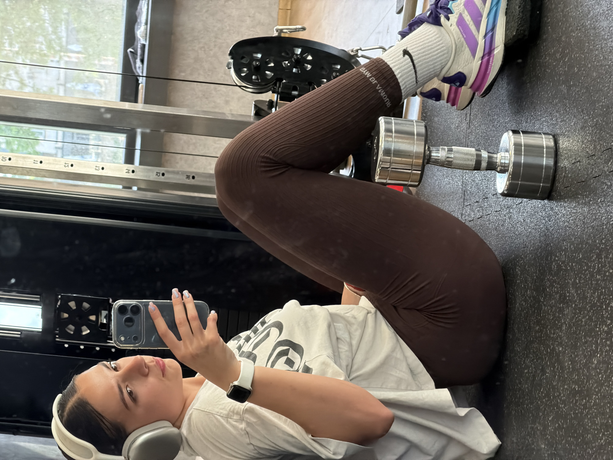 A young woman is in a gym. She is lying on her back on a black rubberized floor. She is holding a smartphone in her left hand and taking a selfie. She is wearing white over-ear headphones, a white t-shirt, brown leggings, and white sneakers with blue and purple accents. Her right leg is bent, with a dumbbell resting on her thigh. A weight machine is visible in the background. The location in the city of Gabrovo, Bulgaria is not evident.