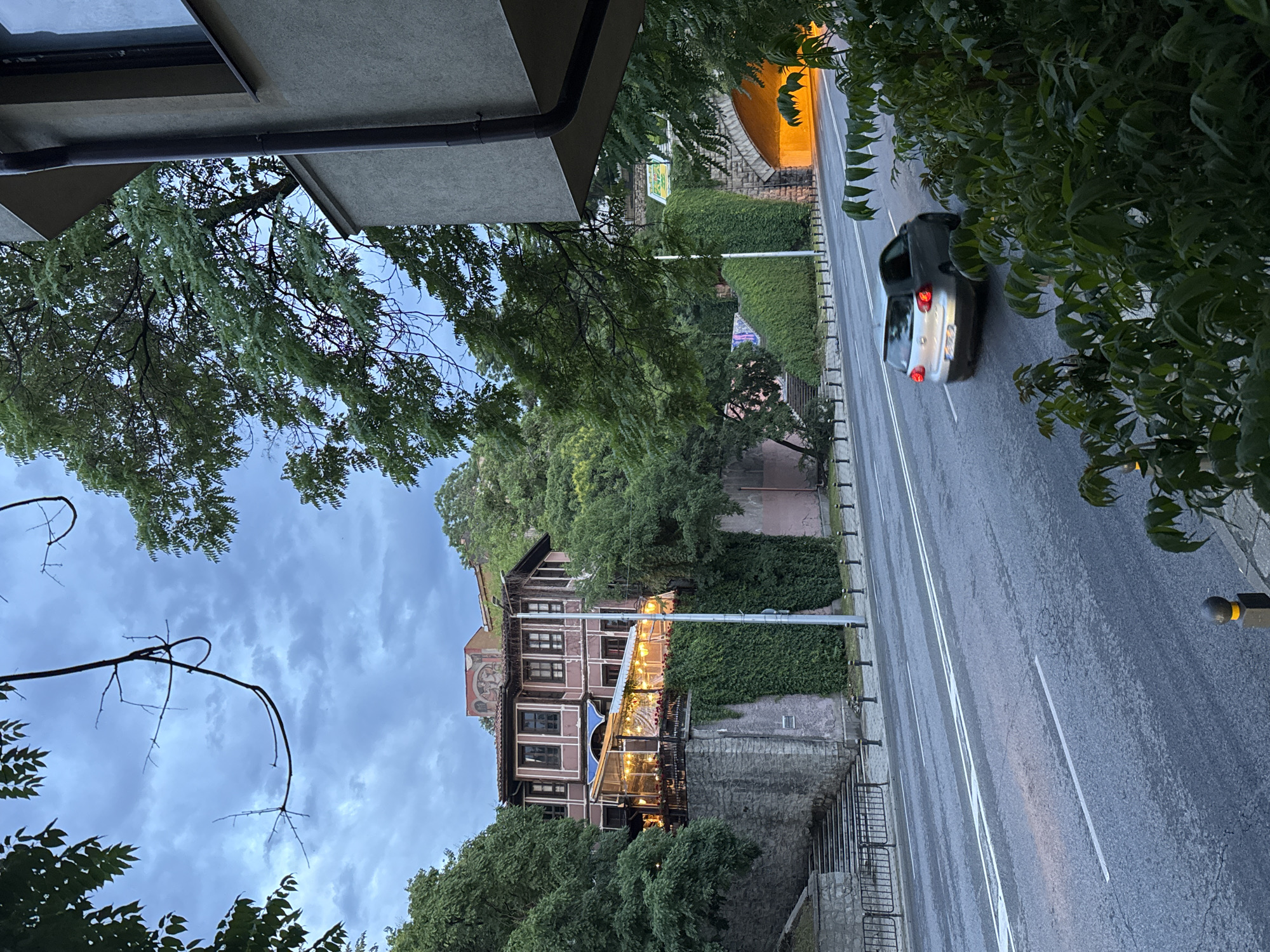 The image presents an elevated, angled view of a street scene in Plovdiv, Bulgaria, captured during dusk or early evening. The sky is overcast with soft blue-grey clouds.
In the midground, a prominent multi-story building showcases traditional Bulgarian Revival architecture, featuring dark timber details, numerous windows, and a faded mural on one section of its facade. The building is partially covered in dense green ivy and hosts an illuminated outdoor seating area, possibly a restaurant or cafe, adorned with numerous string lights that cast a warm, inviting glow. A sturdy stone wall runs along the base of the building, separating it from the street.
A two-lane asphalt road, appearing damp, traverses the scene. White lane markings divide the road, and a single silver/grey sedan is visible driving away from the viewer, its red taillights glowing. The road is bordered by low black bollards and manicured green hedges. Further down the road, a tunnel entrance emits a bright, warm orange light, creating a strong contrast with the cooler ambient light.
The scene is framed by abundant green foliage, with tree branches extending into the foreground and along the sides, partially obscuring the view. No discernible text is visible in the image. The overall atmosphere is quiet and atmospheric, typical of an old town at twilight.