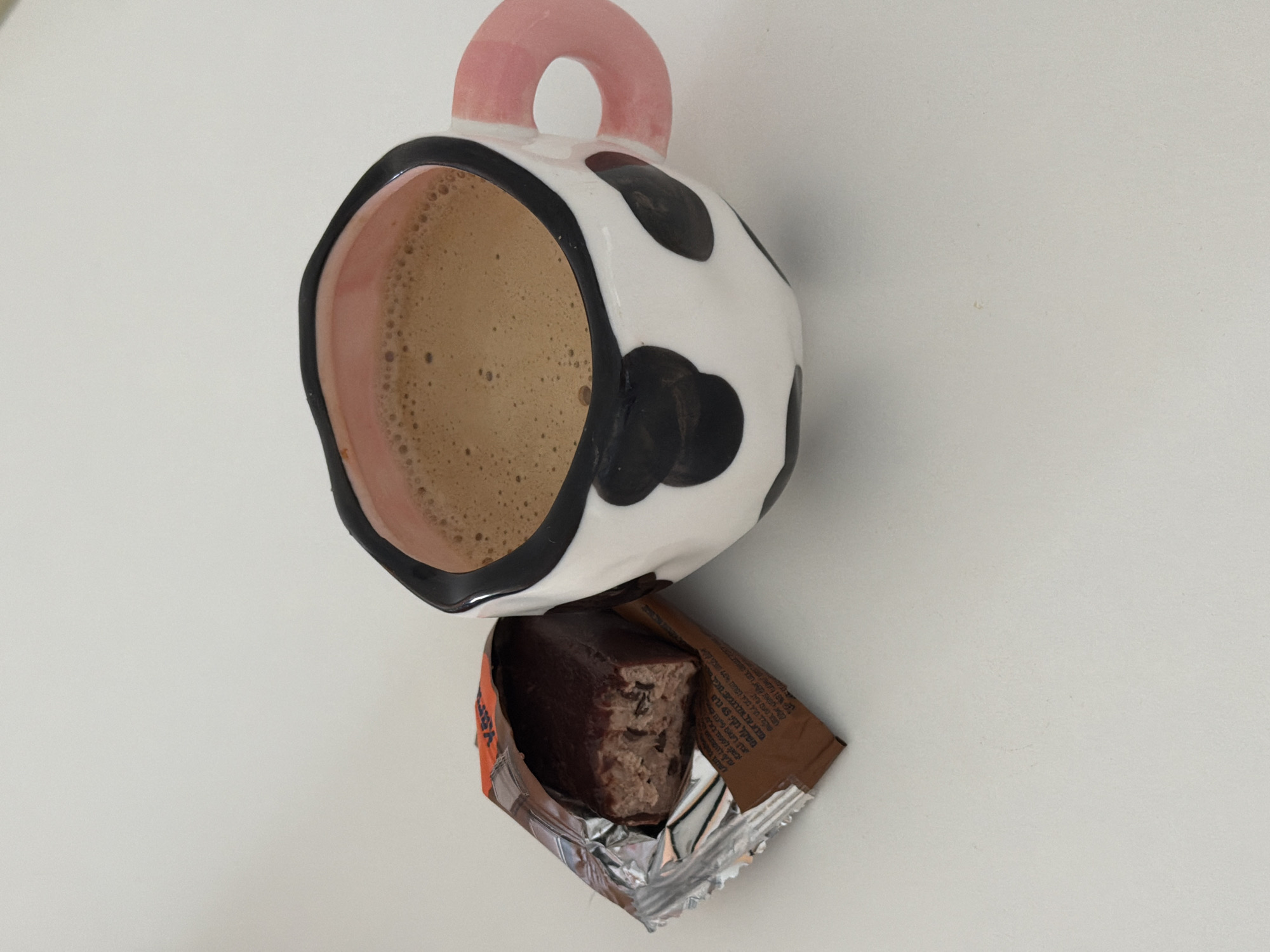 A white ceramic mug with black cow-print spots, a pink interior, and a pink handle is positioned on a plain white surface in Tel-Aviv, Israel. The mug contains a light brown, frothy beverage. To the left of the mug, a chocolate protein bar is partially unwrapped, resting in its brown and silver foil packaging. The bar itself is dark brown with a textured, lighter brown interior visible where it has been exposed, showing inclusions. Hebrew text is visible on the brown portion of the wrapper. No people are present in the scene.
