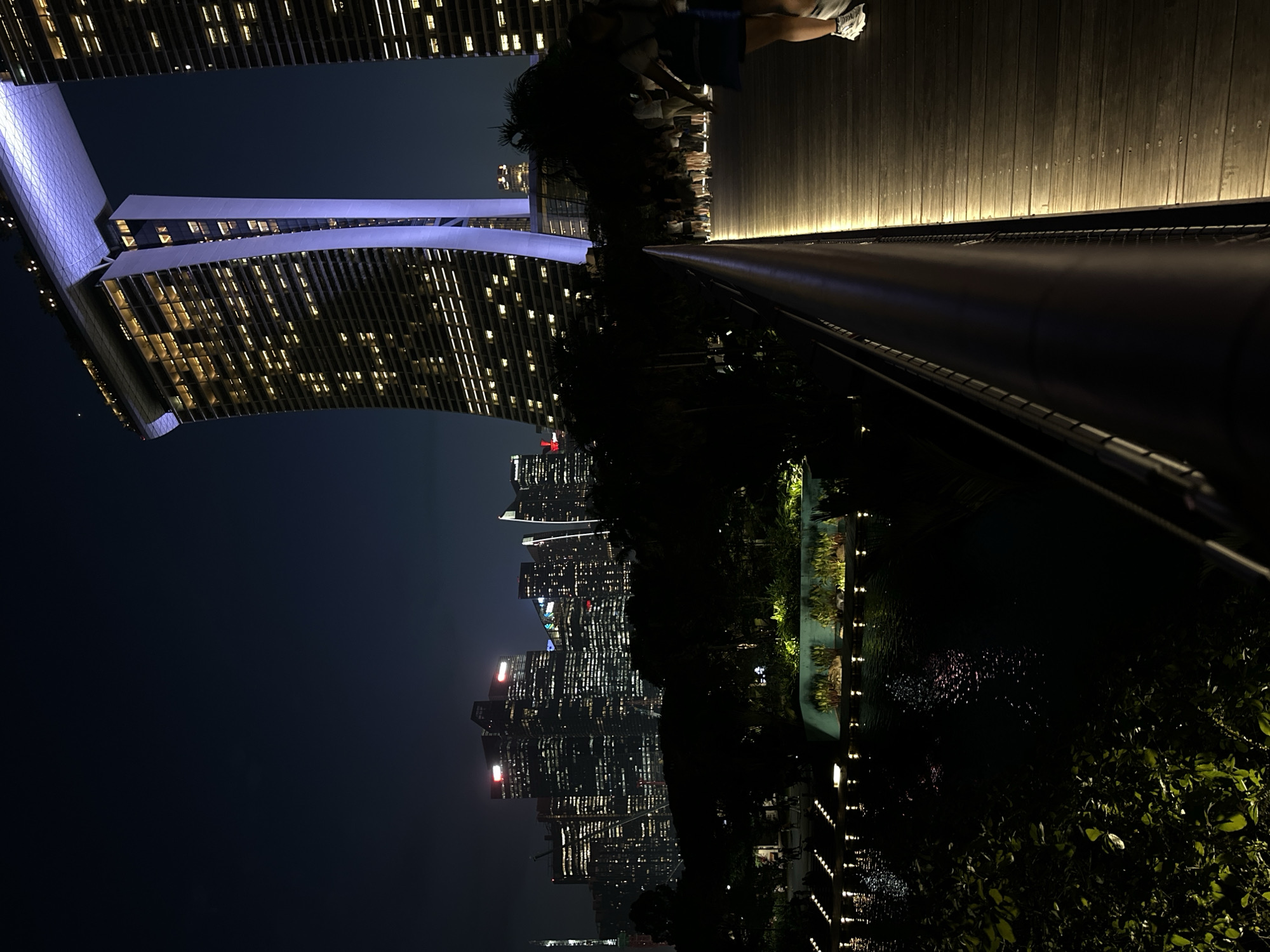 The image captures a nighttime view of Marina Bay Sands in Singapore. A wooden boardwalk, illuminated by string lights, runs along the right side of the frame, leading towards the iconic buildings. Several people are walking on the boardwalk, their figures silhouetted against the lights. In the background, the illuminated skyscrapers of the Singapore skyline rise against the dark night sky. Below the boardwalk and to the left, a body of water reflects the city lights. Lush greenery and planted areas are visible along the water's edge. The overall scene depicts a vibrant urban landscape at night.