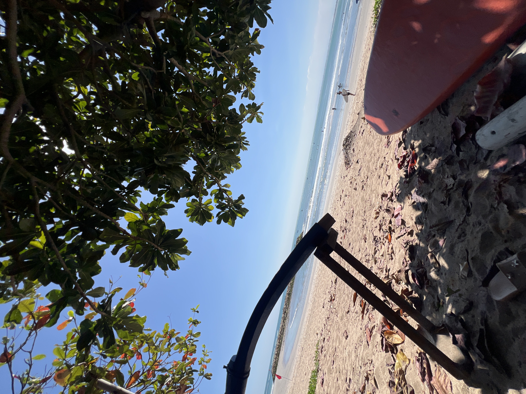 A low-angle shot captures a beach scene in Kuta, Indonesia, under a bright blue sky with dappled sunlight filtering through lush green leaves and branches overhead. In the foreground, the sandy beach is littered with fallen brown leaves. A portion of a red surfboard is visible in the bottom right corner, partially obscured by shadows. To the left, the metal frame of a beach chair or umbrella stand is in focus, casting dark shadows on the sand. Beyond the foreground, the gently lapping waves of the ocean meet the shore. Several surfers are visible in the water and walking on the wet sand near the water's edge. In the distance, a breakwater made of dark rocks extends into the sea, marked by a small red flag. The overall impression is one of a serene tropical beach on a sunny day.