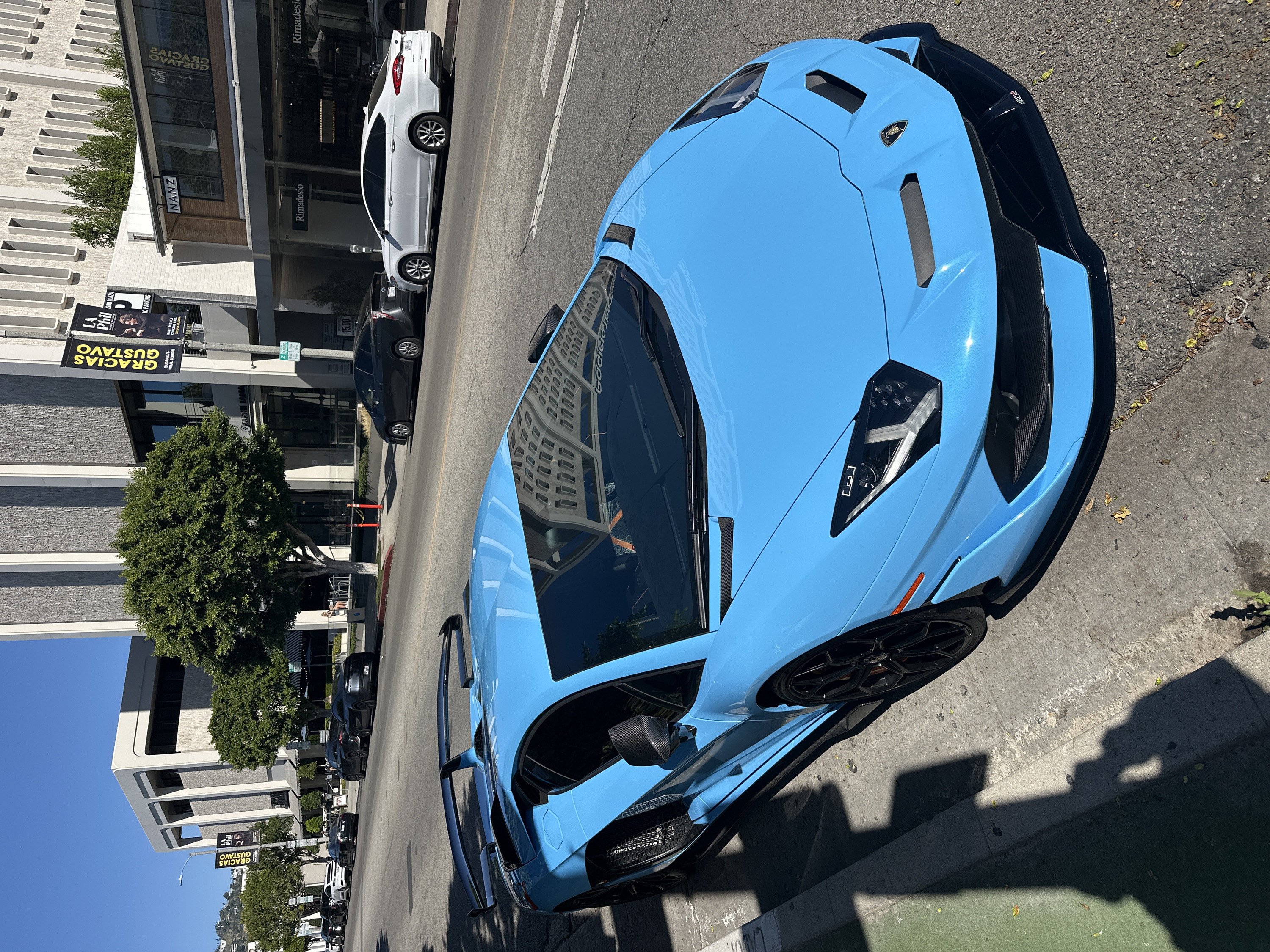 A vibrant blue Lamborghini dominates the frame. The car is parked on the side of a city street with buildings in the background. Banners reading "GRACIAS GUSTAVO" and "LA Phil SON PLAZA" are visible on a lamppost near a tree. "Rimadesio" and "NANZ" are displayed on the storefront. Other parked vehicles, including a white car, line the street, suggesting a busy urban setting. The image is captured from an elevated perspective, showcasing the sleek design of the Lamborghini.