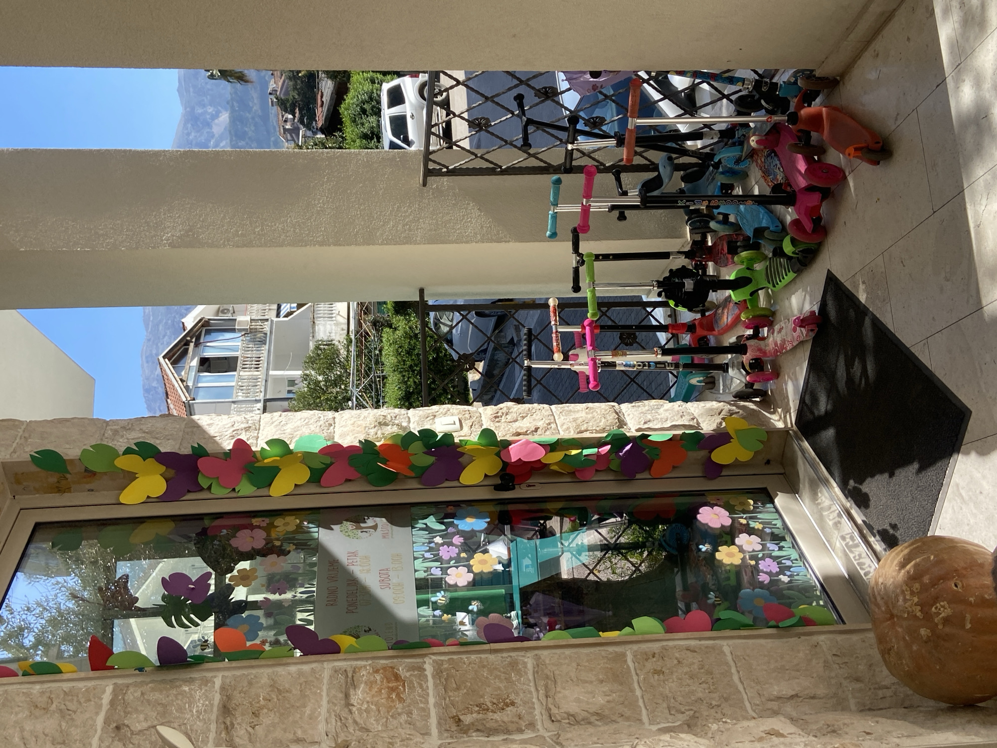 Here is a detailed description of the image:
The image appears to be taken from a sheltered outdoor space, possibly a balcony or porch, overlooking a street or courtyard. The foreground is framed by a light-colored stone or tiled wall and a decorated window. The window is adorned with colorful paper cutouts of flowers, butterflies, and leaves. A round, orange pumpkin sits next to the window frame.
Through the opening, we can see a variety of scooters parked against a metal railing. In the background, buildings and glimpses of a mountain can be seen, indicating an urban or suburban environment with natural surroundings.
The light is bright and the scene appears to be captured on a sunny day. The text "RADNO VRIJEME" is visible on a sign within the window, potentially indicating the opening hours of a business.
The text "TOLEND" and "MASTO Kids Scooter" on the sign indicates this may be a toy store or kids' goods store.
Based on the signs and architecture in the image, this photograph seems to be taken in the city of Budva, Montenegro.