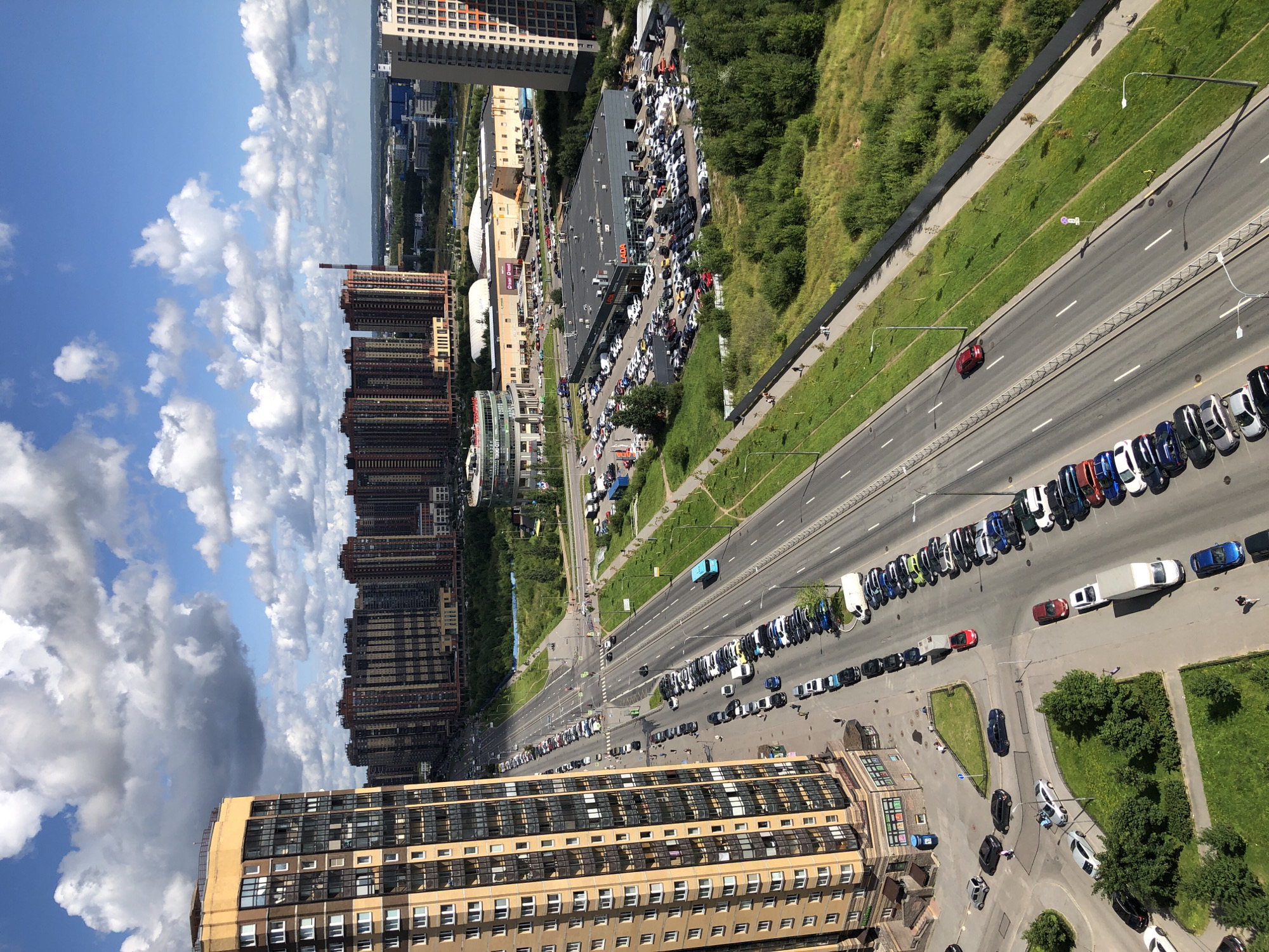 The image shows an aerial view of a city street with high-rise buildings and commercial properties. On the left, a tall, beige apartment building dominates the foreground. Across the street, a multi-lane road is lined with parked cars and a blue bus. Beyond the road, there are several other multi-story buildings, some appearing to be commercial complexes with large parking lots filled with vehicles. A "LADA" branded building is prominently visible among these. Green spaces with trees and grass separate the road from some of the buildings and a fence line. The sky is blue with scattered white clouds. Several individuals are visible walking on sidewalks and in parking areas.