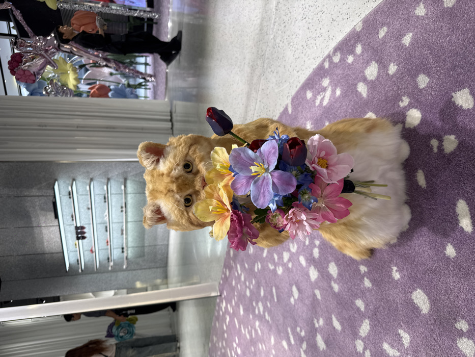 The image displays an interior view of a luxury retail store in Bangkok, Thailand. Centrally positioned in the foreground is a large, plush, ginger and white cat sculpture sitting upright on a lavender-purple carpet with irregular white speckles. The sculpture holds a vibrant bouquet of artificial flowers, featuring yellow, pink, blue, purple, and dark red blossoms, obscuring its lower face.
The carpet transitions to a light-colored, possibly polished concrete or terrazzo floor in the midground and background. To the left, a mirrored surface reflects parts of the store and a person wearing light-colored trousers. Further back on the left, a grey, textured wall features multiple illuminated shelves displaying various eyewear or small accessories. To the right, another individual dressed in dark clothing walks across the light-colored floor, with whimsical, oversized floral and sculptural decorations visible beyond them, suggesting an elaborate display. The environment is brightly lit and appears spacious.