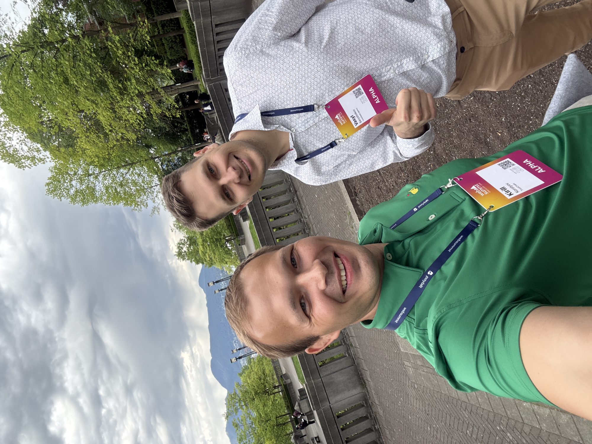 Two men are posing for a selfie outdoors. One man is wearing a white, patterned shirt and khaki pants, and the other is wearing a green polo shirt. Both men are wearing lanyards with badges labeled "Web Summit" and "ALPHA". In the background, there are trees, lamp posts, and a mountain range under a cloudy sky. The location is in Vancouver, Canada. There is also a stone fence.