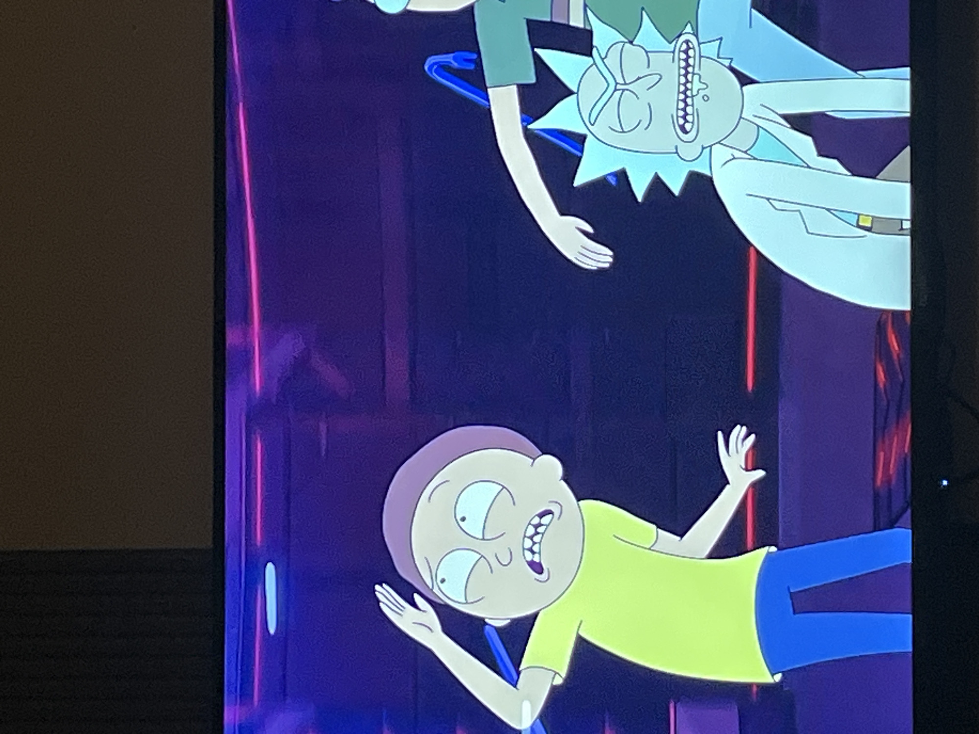 This is a screenshot from the animated television show "Rick and Morty." In the foreground, on the left, is a character resembling Morty Smith, wearing a yellow t-shirt and blue pants, with his hand raised to his forehead and a distressed expression. To the right, and slightly behind, is a character resembling Rick Sanchez, wearing a lab coat over a teal shirt and blue pants, with his head tilted down and a manic grin. The background is a dark, futuristic setting with purple and pink neon lighting. The image appears to be taken from a television screen, likely in a domestic setting. There is no discernible location in the city of Krasnoyarsk, Russia, present in this image.