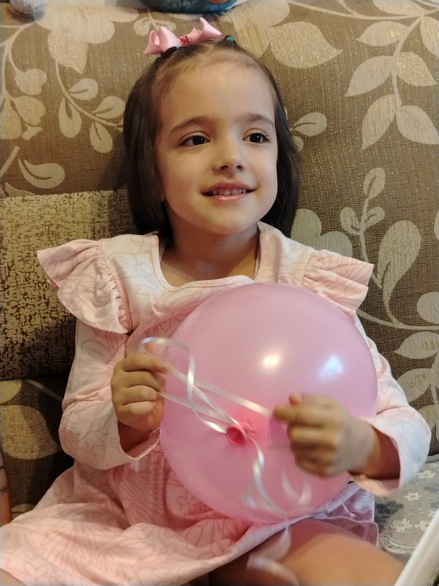 A young girl is sitting indoors, presumably on a sofa, holding a pink balloon with white ribbons. She has dark hair with a pink bow and is wearing a light pink dress with ruffled sleeves. The background is a patterned fabric, likely the sofa's upholstery. There is no indication of location in the city of Noviy, Russia.