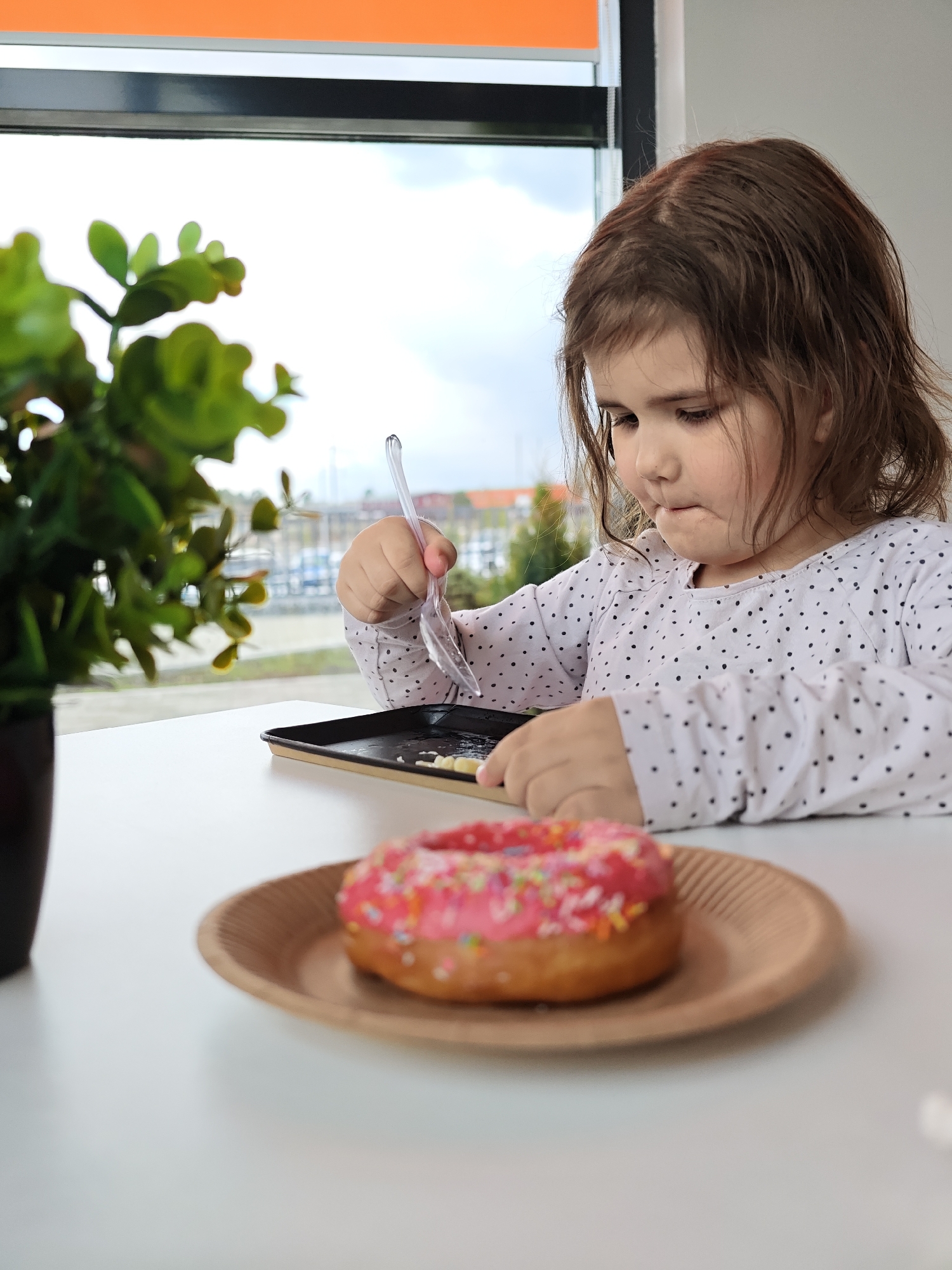 The image features a candid, close-up shot of a young child at a table, intensely focused on a task. The child, with medium-brown hair, is wearing a white long-sleeved shirt adorned with small black polka dots. They are holding a clear plastic spoon over a small black rectangular tray, which appears to contain some food residue or crumbs. The child's mouth is slightly pursed, conveying concentration or a thoughtful expression.
In the foreground, a vibrant pink donut, covered in colorful sprinkles, sits on a round, light brown plate on the white table. To the left, a green potted plant is visible, partially out of focus. The setting appears to be indoors, with a large window serving as the background.
Through the window, an overcast sky is visible, suggesting daytime. Blurred buildings and structures can be discerned in the distance, indicating an urban or town environment, aligning with the location context of Votkinsk, Russia. An orange element, possibly part of an awning or interior frame, is visible at the very top of the window. The overall scene captures a quiet moment of a child enjoying a snack or engaging with their food. No visible text is present in the image.