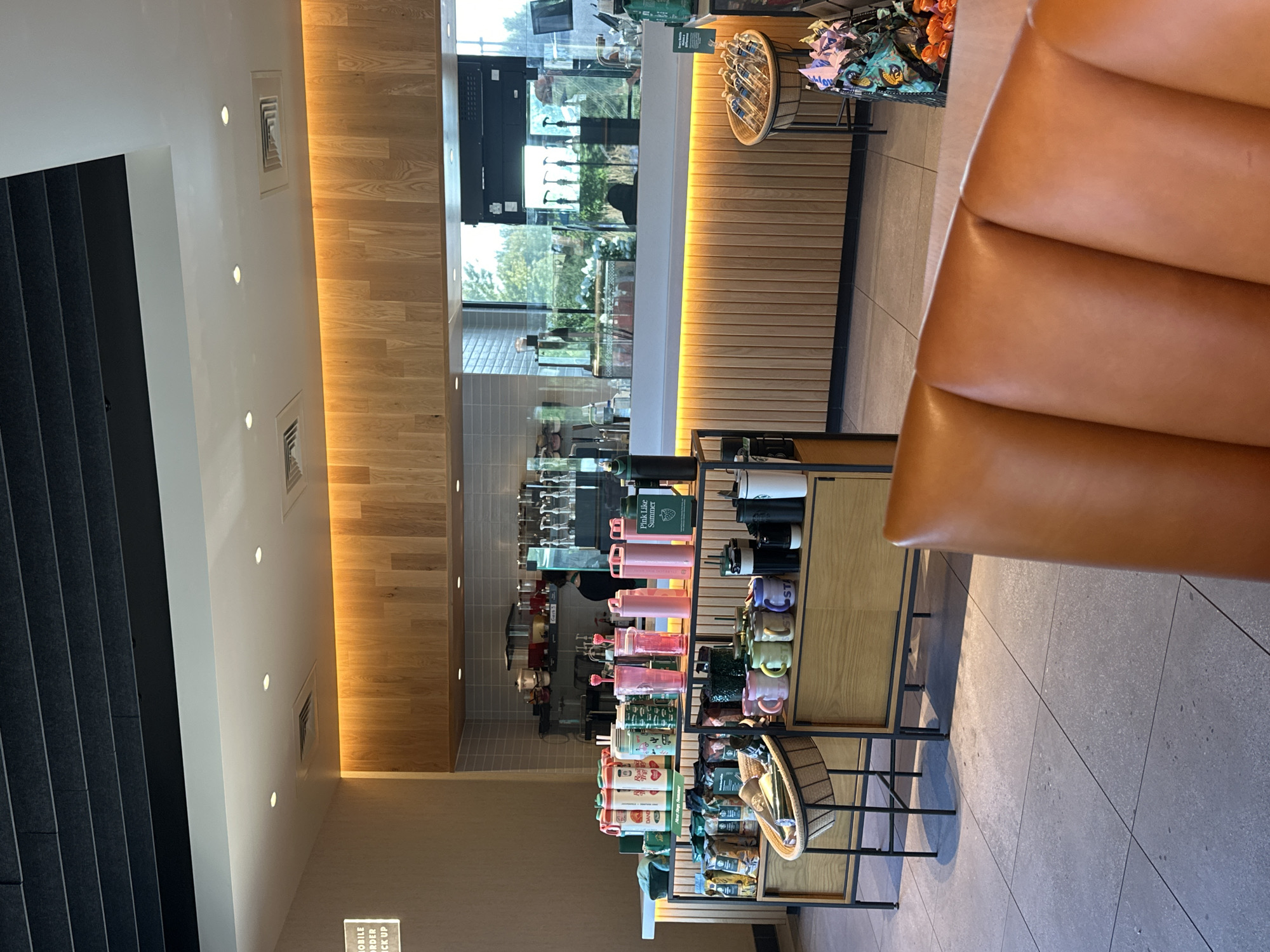 This is an indoor image of a Starbucks store in McHenry, United States of America. The perspective is from a seated position, looking towards a counter with merchandise. The counter is made of light-colored wood and is stocked with various Starbucks branded items, including colorful tumblers, mugs, and packaged coffee. A basket filled with bottles is visible on the right side of the counter, and a rack with a variety of bags of coffee is on the left. Behind the counter, a barista is partially visible, and behind them is a window offering a view of greenery. The ceiling has a dark, ribbed paneling and recessed lighting. The floor is tiled with large, gray tiles. A plush, orange leather booth seat is visible in the foreground, partially obscuring the view.