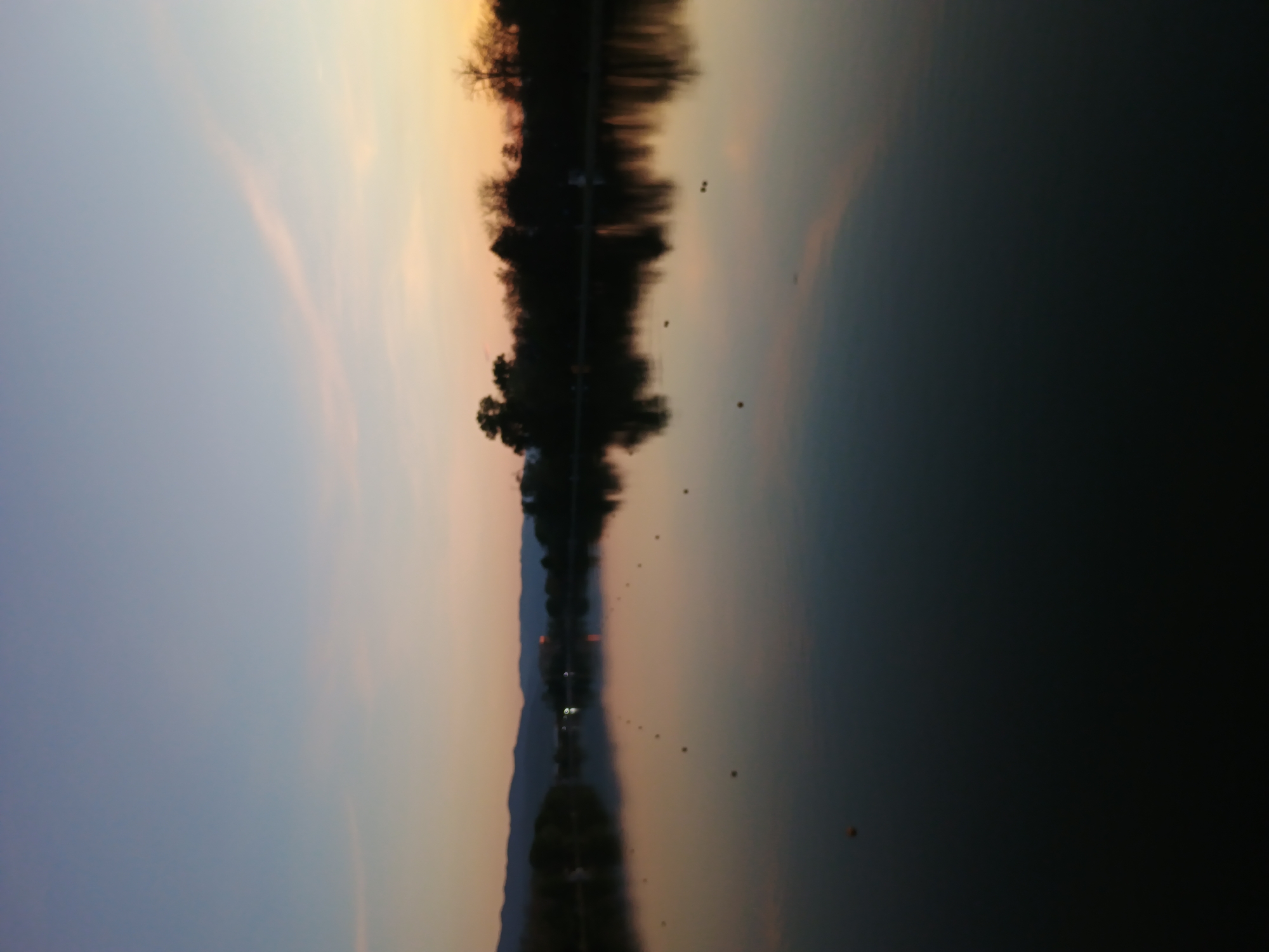 A serene landscape photograph captures a tranquil body of water under a sky painted with the soft hues of dawn or dusk. The image is oriented with the sky occupying the upper portion, transitioning from gentle blue and purple tones on the left to warm orange and yellow shades on the right, suggesting either sunrise or sunset.
A dark, silhouetted strip of land, likely a distant shoreline lined with trees and possibly some low-lying mountains or structures, stretches horizontally across the middle of the frame, separating the sky from the water. This landmass and its flora are reflected almost perfectly in the calm water below, creating a symmetrical visual effect.
The lower half of the image is dominated by the still water, which acts as a mirror, faithfully reflecting the colorful sky and the dark, silhouetted landscape. Scattered across the water's surface are several small, dark, round objects, which could be buoys marking a course or distant waterfowl like ducks or geese.
The scene conveys a sense of peace and stillness. The clear conditions and soft lighting contribute to the tranquil atmosphere. There are no visible people or explicit activities taking place, and no discernable text. Given the context, this picturesque view is likely from Plovdiv, Bulgaria, possibly along the Maritsa River or one of the city's parks featuring a body of water.