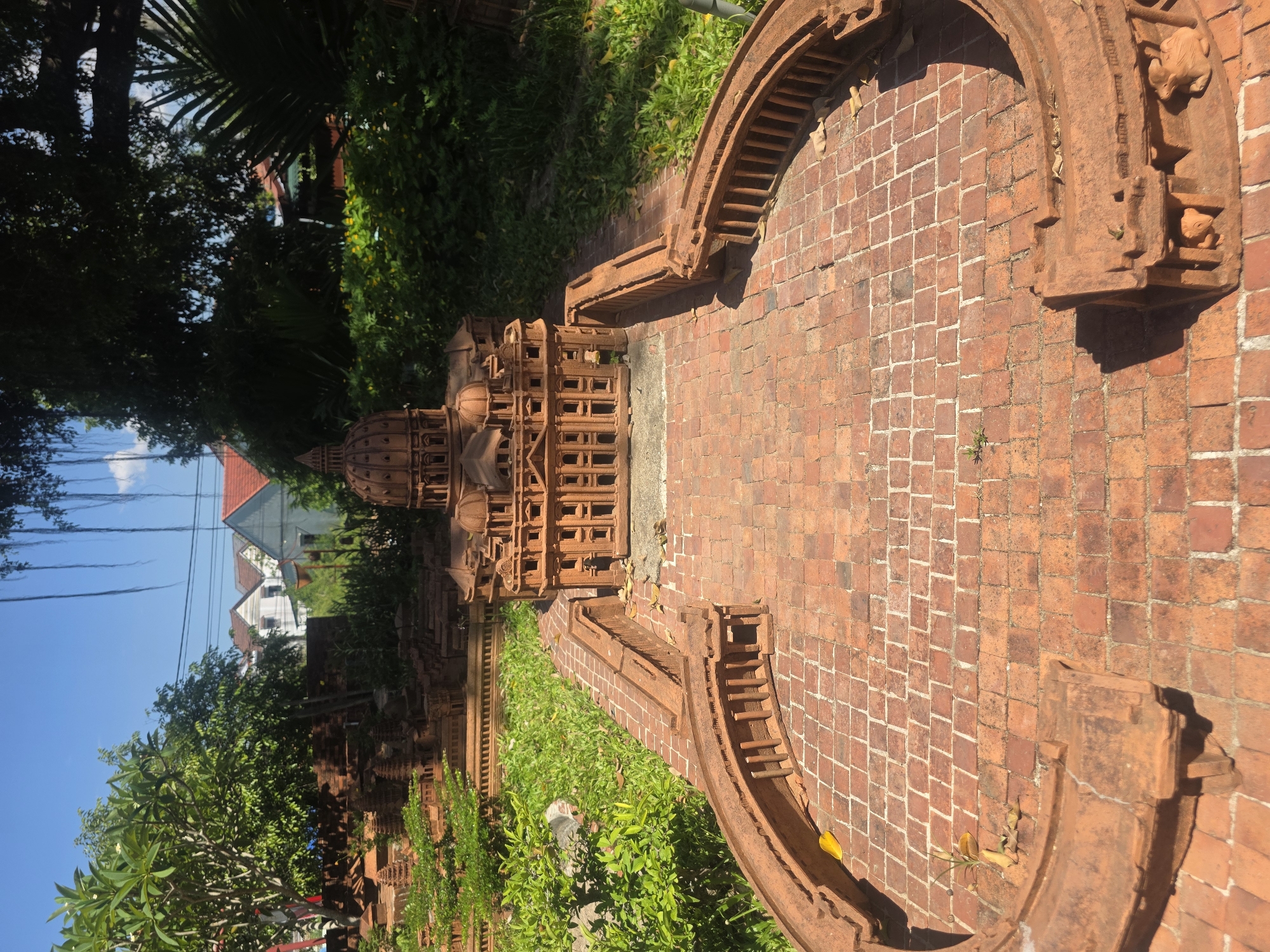 The image captures an outdoor scene featuring an intricate display of miniature architectural models crafted from reddish-brown brick or terracotta. A prominent model, resembling a multi-tiered temple or pagoda with a large central dome and detailed, stacked levels, stands on a red brick pathway. This pathway is composed of square bricks laid in both straight and diagonal patterns.
Adjacent to the models, decorative arched structures made of similar reddish brick frame sections of the pathway. One such arch includes small carved figures, possibly depicting animals like frogs, integrated into its design. The models and pathway are surrounded by lush green grass and various trees, including tall palm fronds and other leafy foliage, suggesting a tropical environment. In the background, under a bright blue sky dotted with white clouds, several residential houses with red roofs and white walls are visible amongst the trees.
The scene is well-lit by natural sunlight, casting distinct shadows, indicating it is daytime, likely morning or afternoon. No people are visible, and no specific activity is taking place beyond the static display of the miniature structures. The tropical vegetation and architectural style align with the given location context of Đà Nẵng, Vietnam. No discernible text is visible in the image.