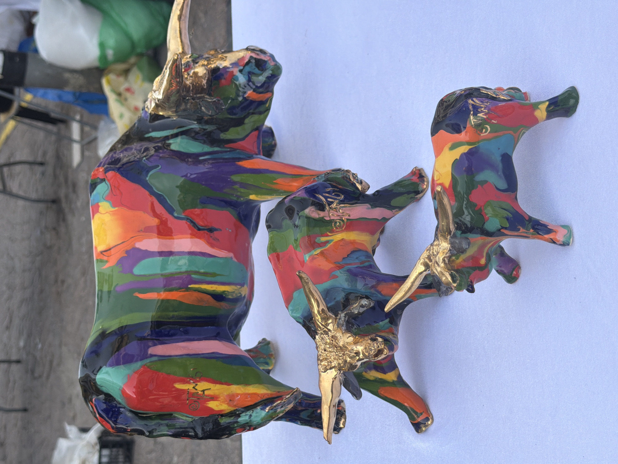 Two brightly colored, abstract sculptures of bovine figures, likely bulls or oxen, are depicted against a plain white background. The original image is rotated 90 degrees clockwise, so the sculptures appear to stand upright on the white surface.
The larger sculpture, positioned on the left, and a smaller one, on the right, are coated in a thick, glossy, multi-colored paint. The colors swirl and blend across their bodies, resembling melted crayons or poured enamel, with prominent shades of red, orange, yellow, green, blue, purple, pink, and black. Both sculptures feature metallic, golden horns and ears, which contrast with their vibrant bodies. The golden elements appear to have a textured, slightly rough finish.
Beyond the white background, an out-of-focus outdoor setting is visible. This blurry background shows concrete or paved ground, indistinct metal structures (possibly part of a cart or railing), and several blurred objects, including white and green bags or refuse. This suggests an urban outdoor environment, possibly a street vendor's display or market stall in Tel-Aviv, Israel.
No specific activity is taking place beyond the display of these art pieces. The lighting indicates bright daylight.
Notable details include handwritten-like markings or signatures in gold on the sculptures. On the lower side of the larger bull, a gold inscription resembling "OMYG" or a similar signature is visible. On the head/neck area of the smaller bull, another gold marking, possibly "OMY" or a similar set of initials, can be seen. The overall aesthetic is one of modern, vibrant folk art or contemporary sculpture.