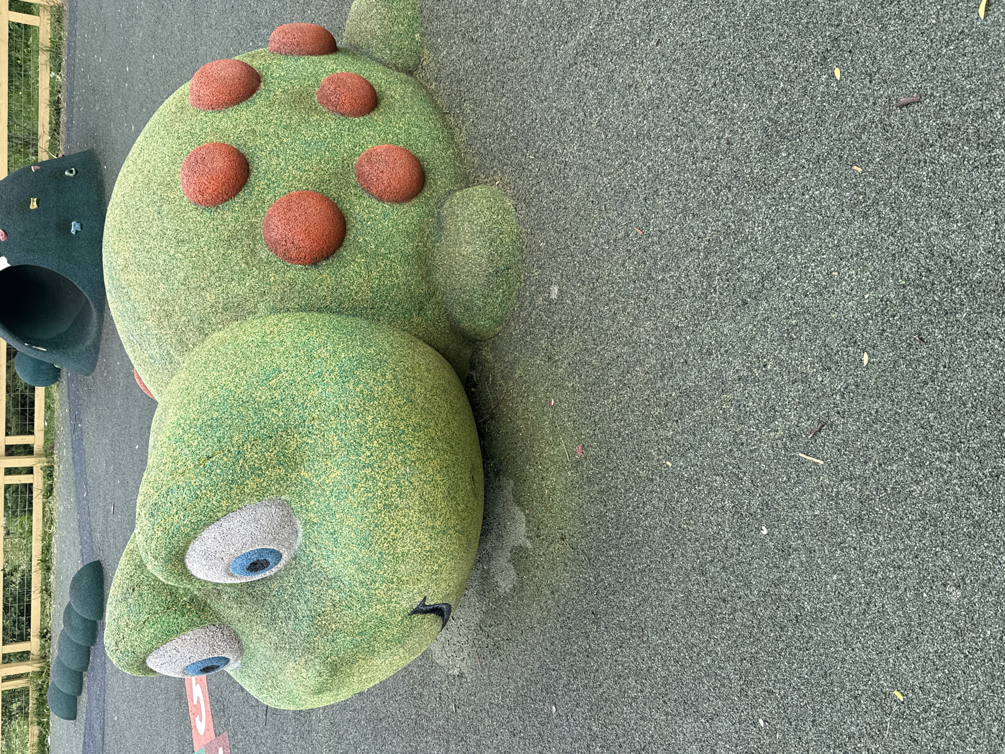 The image depicts a playground in Dmitrov, Russia. The main feature is a large, green, stylized turtle sculpture with red spots on its shell and blue eyes. The turtle appears to be made of a textured material, possibly a soft, rubberized surface for safety. To the left of the turtle, there are sections of a climbing structure and a tunnel. The ground is covered in a gray-green, granulated safety surface. A fence is visible in the background on the left. The scene appears to be empty of people.
