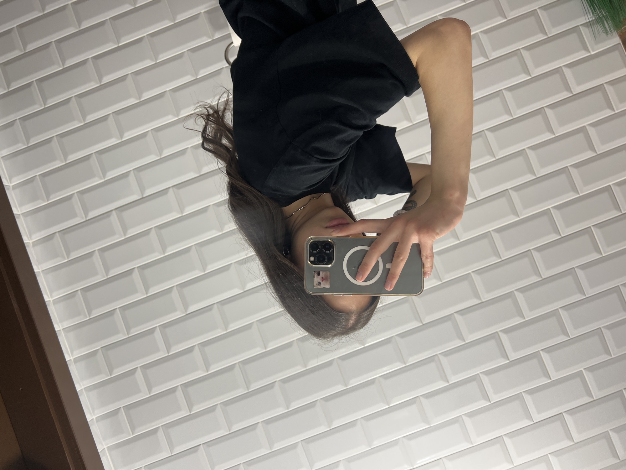 A mirrored image shows a person holding a smartphone in front of a tiled wall. The person is wearing a black T-shirt and a silver necklace. The smartphone has a grey case with a MagSafe ring and a sticker with a cat image. The tiled wall is white with a brick pattern. The environment appears to be a bathroom or similar space with reflective surfaces. The location, Ternopil, Ukraine, is not deducible from the image.