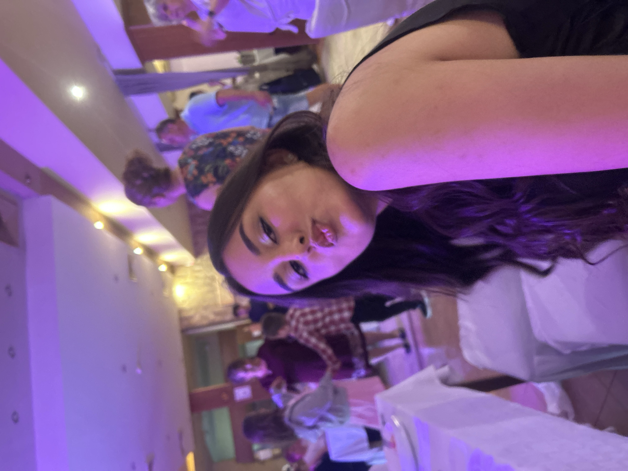 The image shows a young woman taking a selfie in a dimly lit indoor environment. She is making a pouting face towards the camera. The background reveals what appears to be a restaurant or banquet hall setting, with tables covered in white tablecloths and other people milling around in the background. The lighting is dominated by a strong purple hue. Details about the city of Elhovo, Bulgaria, cannot be definitively verified from the image.