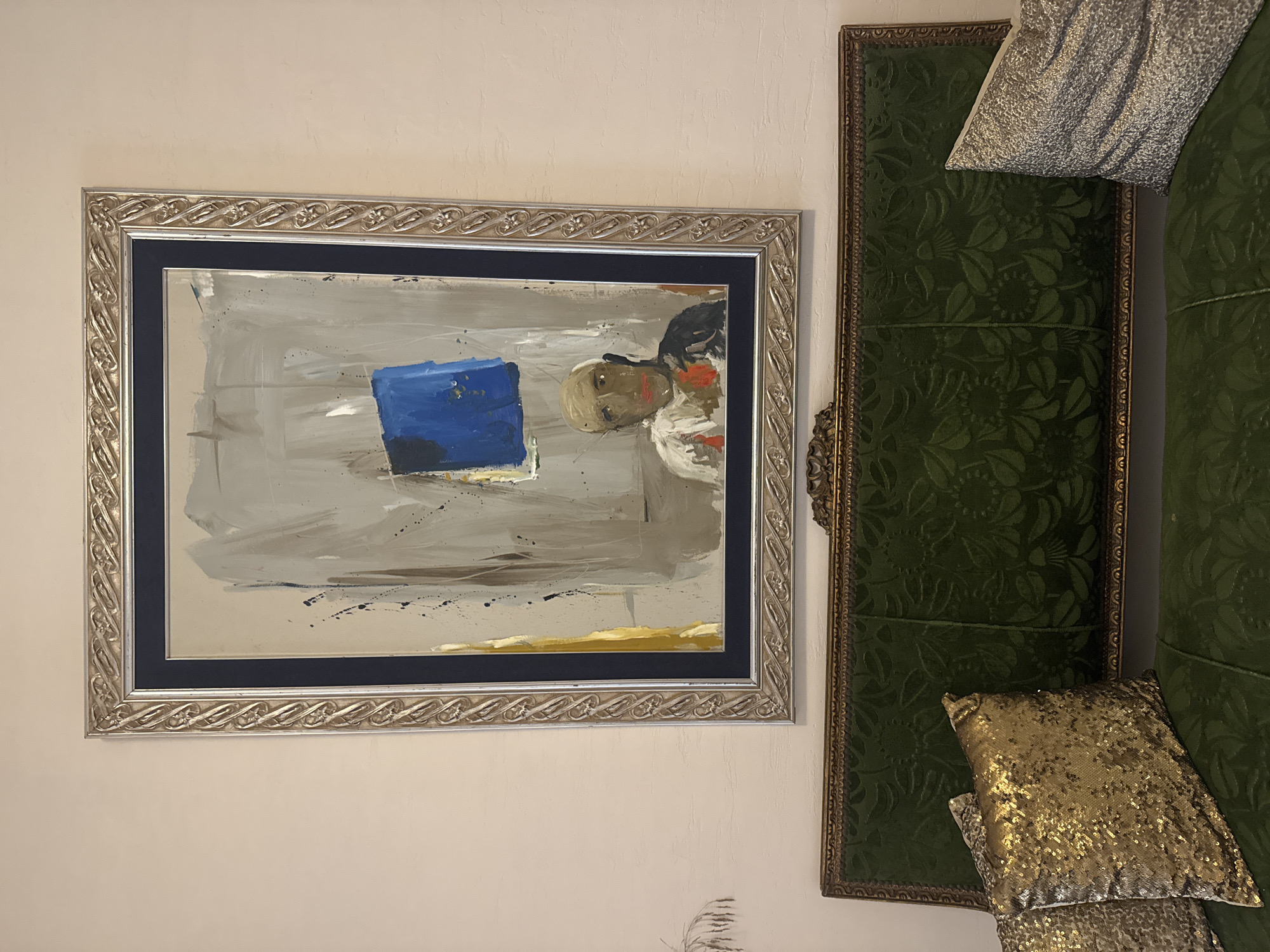 The image shows a framed abstract painting hanging on a plain, light-colored wall above a dark green upholstered sofa. The painting features a man's face with a blue square above it, rendered in broad strokes of gray, white, and black paint, with splashes of yellow and red. The frame of the painting is ornate and silver-toned. The sofa has a textured, patterned upholstery and is adorned with two decorative pillows: one covered in gold sequins and another in a silver, textured fabric. The setting appears to be an interior space, possibly a living room, in an unknown location.