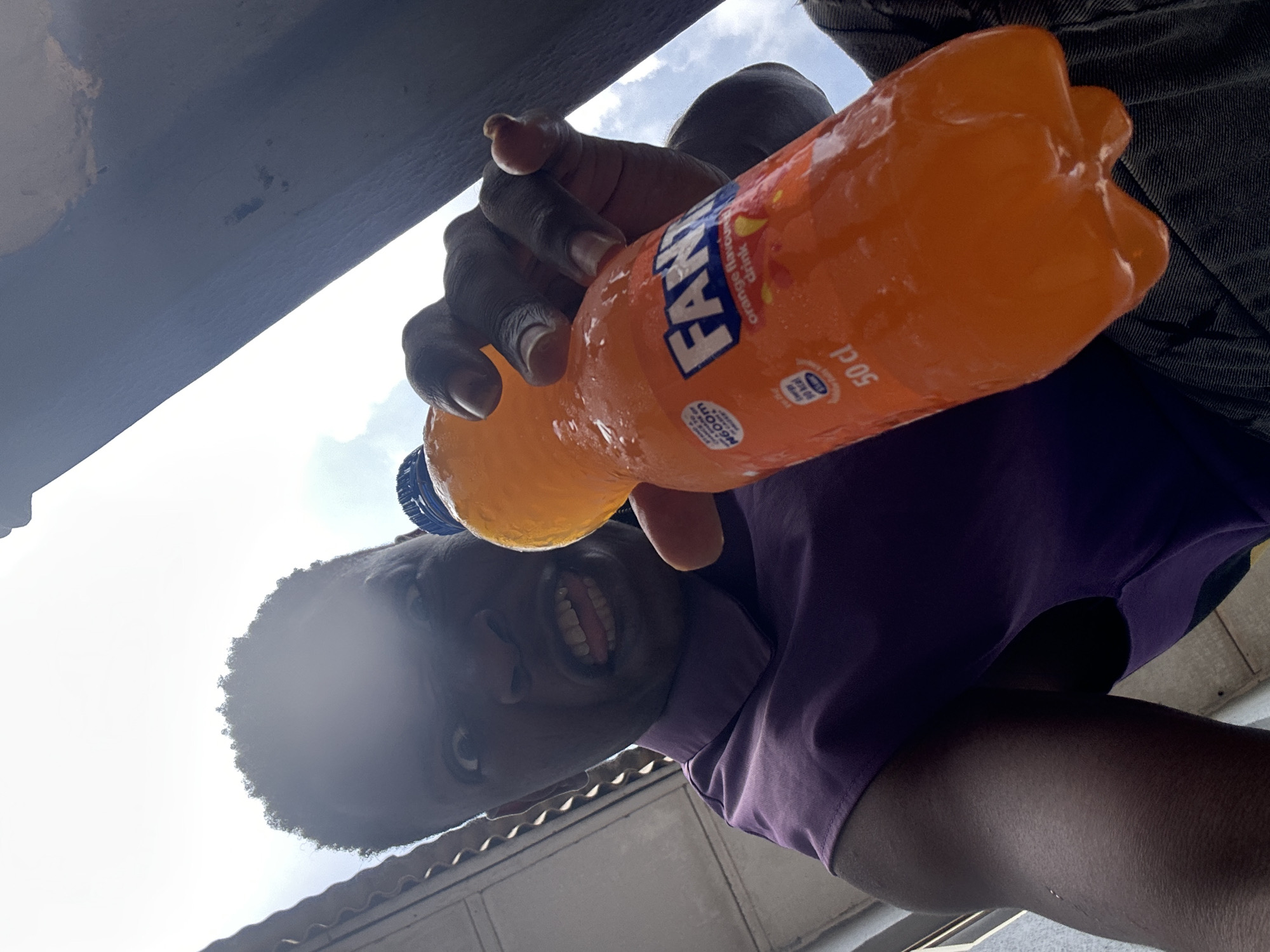 A low-angle shot features a person holding a bottle of Fanta Orange in their right hand. The bottle appears to be chilled, with condensation visible on its surface. The person is wearing a purple shirt and jeans. The background includes a cloudy sky and what appears to be the underside of a roof or overhang. The image suggests an outdoor location. There is no evidence of the Ikorodu environment in the image.