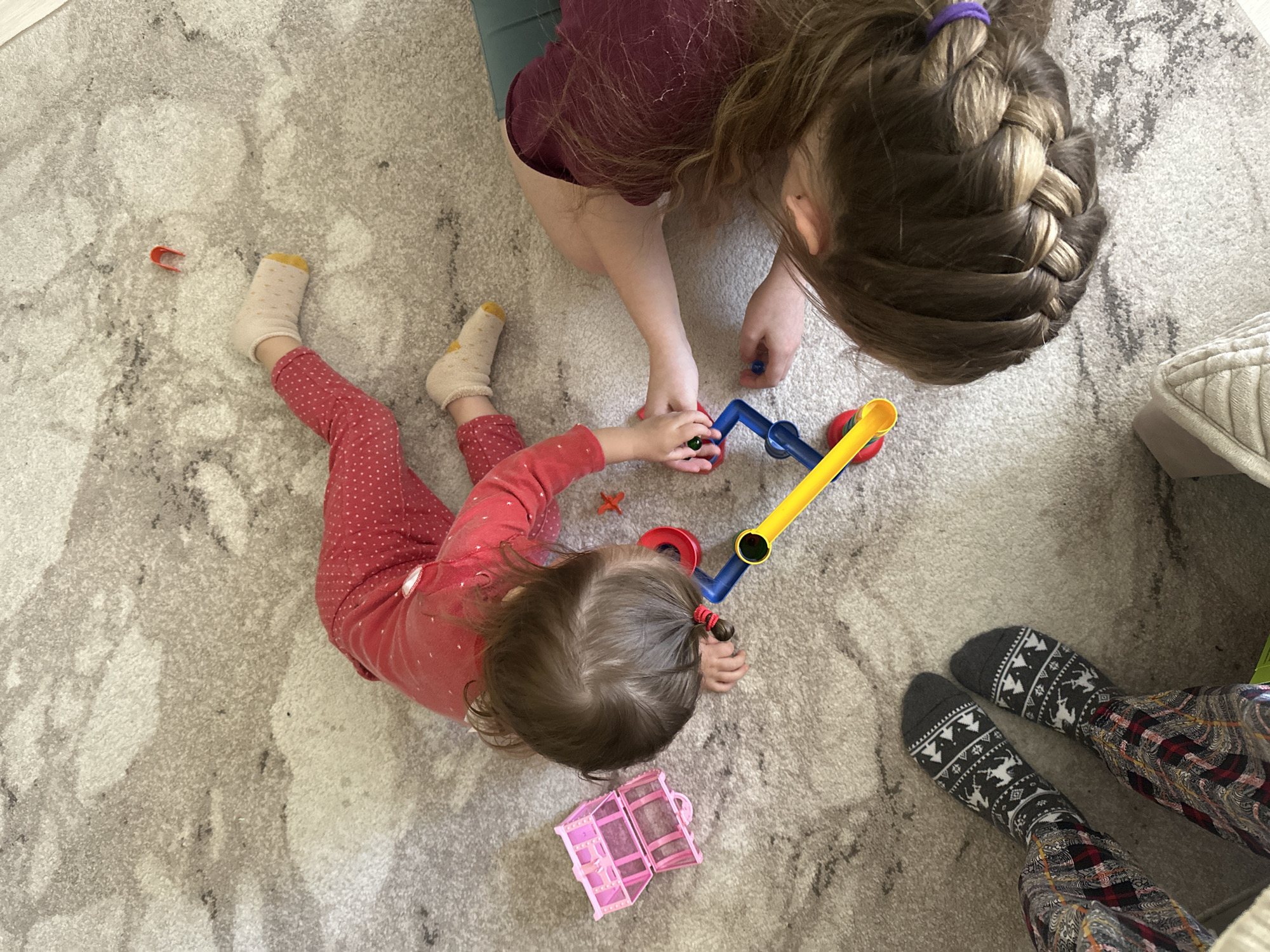 This overhead image captures an indoor scene in Uva, Russia, featuring two children playing on a light-colored, patterned carpet. An older child, with long brown hair styled in a French braid and secured with a purple elastic, kneels in the upper portion of the frame, wearing a burgundy top and teal trousers. They are seen interacting with a younger child, who lies on their stomach in the foreground. This younger child wears a red polka-dot onesie or top and pants, cream-colored socks with yellow toes, and has short brown hair tied in small pigtails.
A colorful plastic push toy, primarily yellow and blue with red elements, is central to their play, with both children's hands near it. A small, open pink plastic toy resembling a castle or house lies near the younger child, along with a tiny orange plastic piece. In the bottom right corner, the feet of a third individual are visible, clad in grey patterned socks with white winter motifs (trees and deer) and plaid trousers, resting next to a light-colored upholstered cushion. The scene suggests a relaxed and playful interaction between the children during what appears to be daytime. No text is visible within the image.