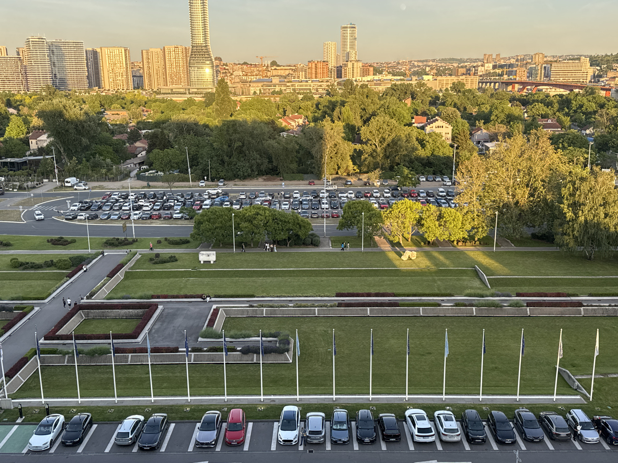 The image shows an elevated view of a parking lot, flags, lawn, gardens, and city buildings in Unknown, Unknown.
In the foreground, a row of parked cars aligns with a line of flagpoles. The flagpoles run along the edge of a green lawn divided into rectangular sections by walkways. The lawn is flanked by landscaped areas with bushes and flower beds. Pedestrians can be seen walking along the walkways.
In the midground, a large parking lot filled with vehicles is visible. Beyond the parking lot is a dense area of trees and buildings, likely residential areas.
In the background, the city skyline features tall, modern buildings, including what appear to be residential skyscrapers. A bridge structure is also visible in the distance.
The image is taken in daylight, with a clear sky and soft lighting. The scene appears to be taken at the Serbian Palace in Belgrade.