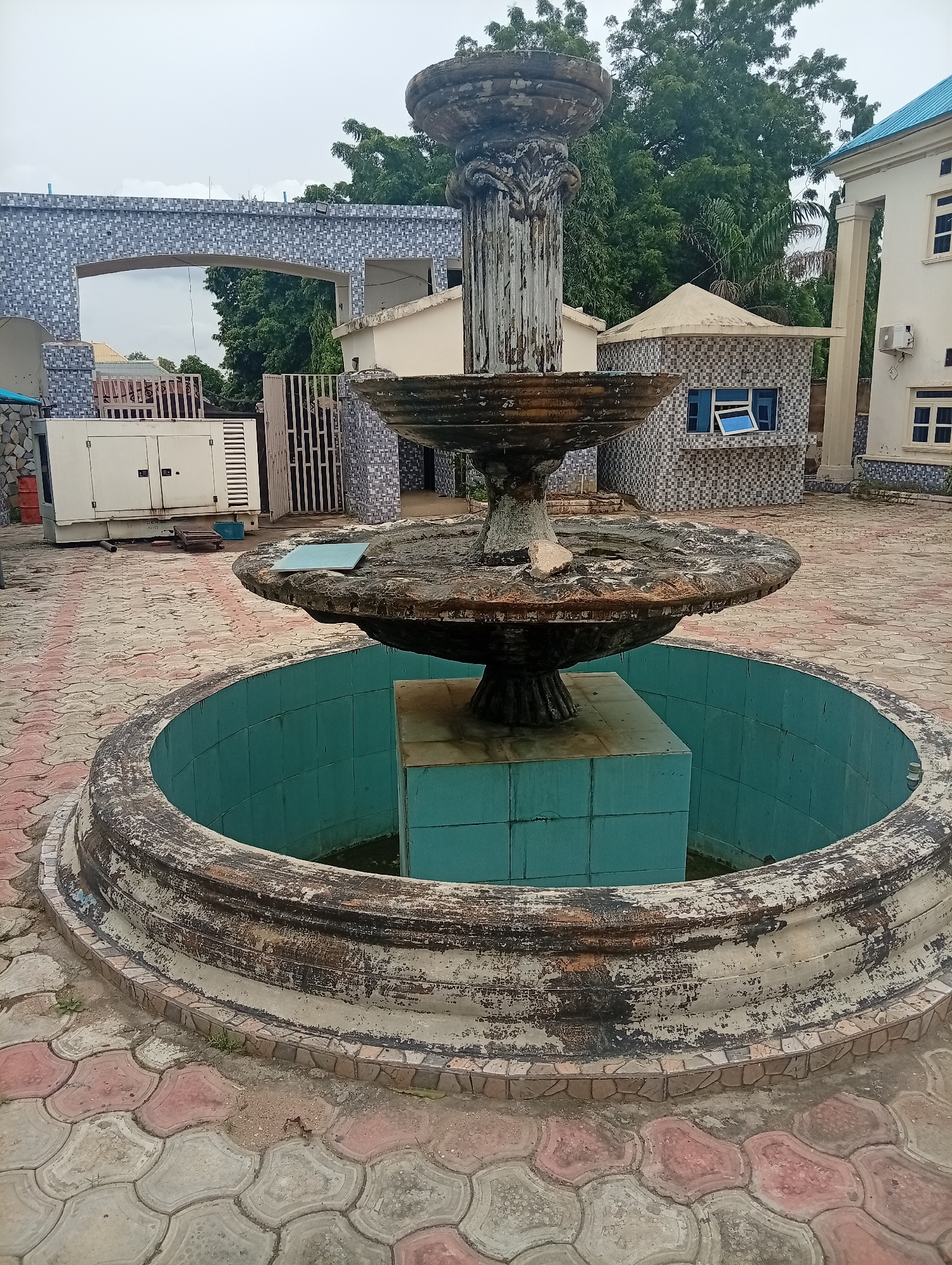 This media file depicts an outdoor scene in Jalingo, Nigeria. The primary subject is a weathered, three-tiered fountain situated in a paved courtyard. The fountain appears to be made of concrete and shows signs of age, with peeling paint and debris. The base of the fountain is a circular basin tiled in a vibrant turquoise color, with some water visible at the bottom.
Behind the fountain, the setting includes architectural elements such as an arched entryway with intricate blue and white mosaic tiles, a white generator, and a small building with similar mosaic tiling and blue-framed windows. To the right, a light-colored building with a blue roof and multiple windows is visible, suggesting it might be a residential or commercial structure. Lush green trees form a backdrop, indicating a potentially warm climate.
The ground is paved with interlocking concrete blocks in a pattern of red and grey. The sky is overcast, suggesting it is either early morning or late afternoon, and the weather appears to be cloudy. No people are visible in the scene, and no specific activity is taking place. The overall impression is one of a functional but somewhat neglected public or private space. No visible text is present in the image.