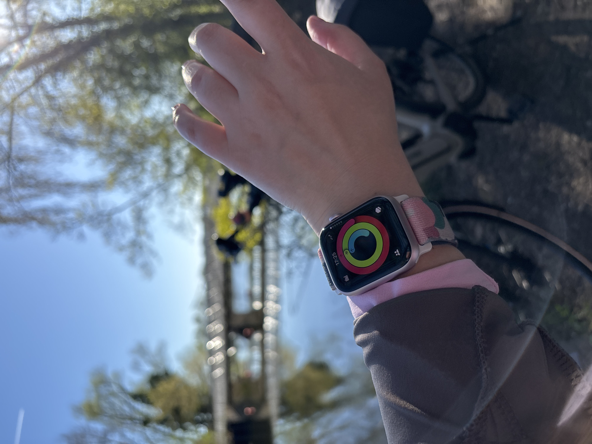 A person's left hand and wrist are in focus, showing an Apple Watch with activity rings displayed on the screen. The watch has a patterned wristband with pink and green hues. A pink sleeve peeks out from underneath a gray long-sleeved shirt. The background is blurred, revealing a low-angle view of tall trees and a blue sky. A bicycle frame is partially visible to the right of the wrist. An elevated structure is visible through the trees. The location is Unknown, Unknown.