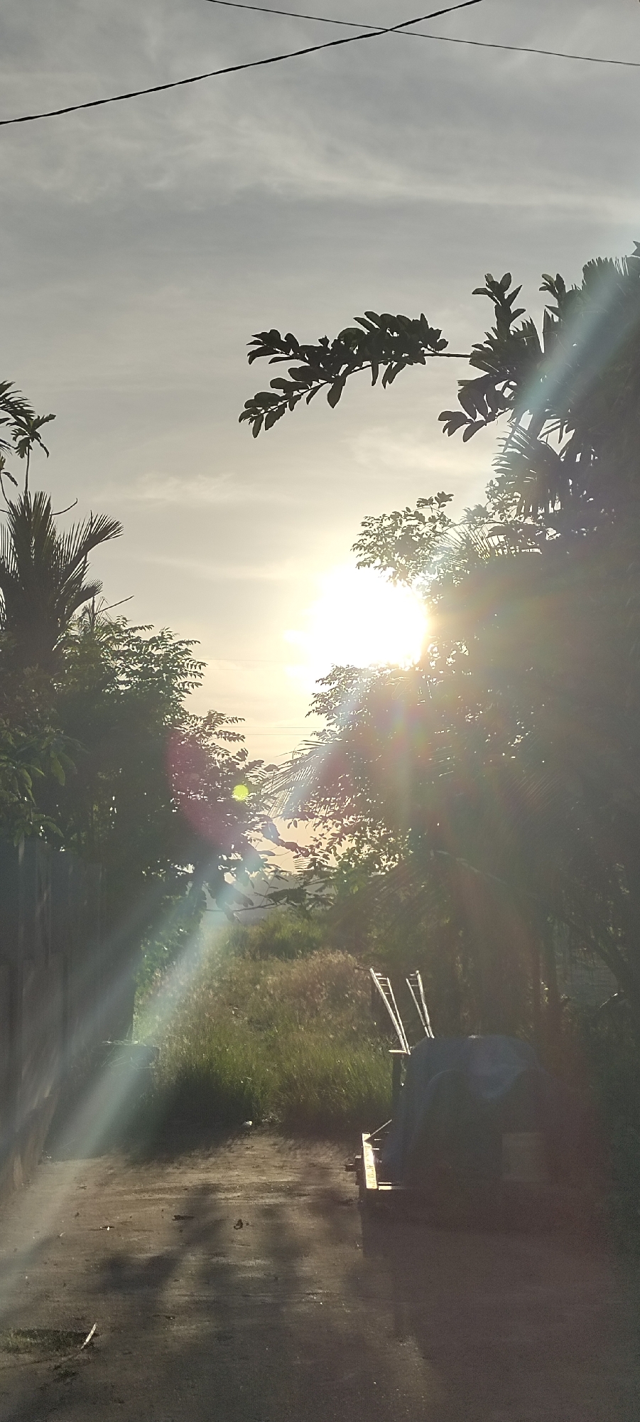 The image shows a sunlit scene in Lhokseumawe, Indonesia. The sun is low in the sky, creating intense glare and lens flares, partially obscured by trees. A dirt road or path is visible in the foreground, leading towards the light. A structure covered by a tarp is positioned on the right side of the road with ladders. There is greenery on both sides of the path, with trees and tall grass. Power lines are seen overhead. The sky is bright, suggesting either sunrise or sunset.