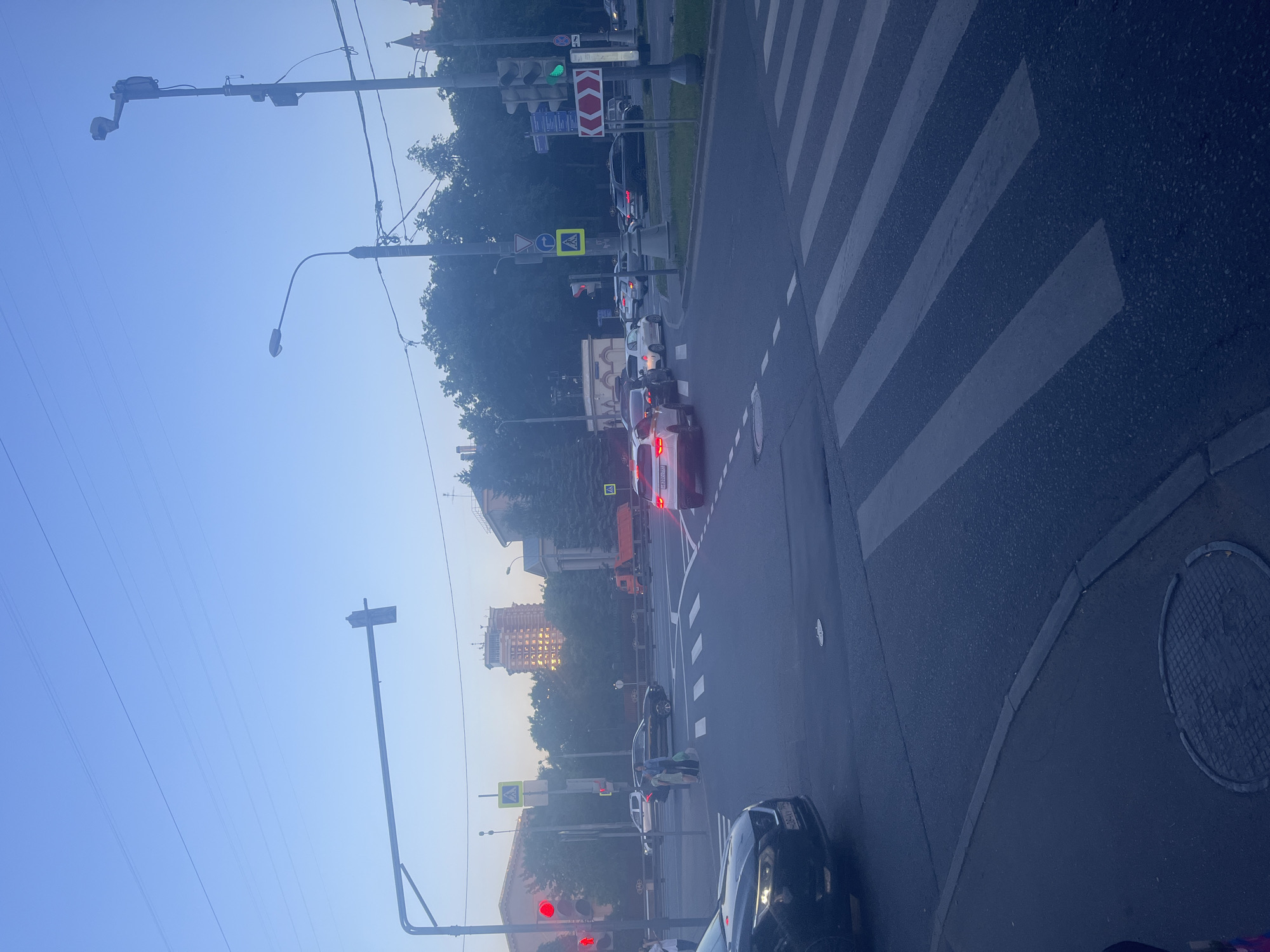 An overpass intersection in Khimki, Russia, is depicted during dusk, bathed in the soft glow of streetlights and the fading daylight. A pedestrian crossing is visible in the foreground, with cars navigating the intersection. A lone figure stands on the sidewalk, observing the traffic. In the background, a towering building with illuminated windows stands tall against the darkening sky, suggesting an urban setting. The scene is a typical depiction of city life in Russia, with its bustling streets and architectural landmarks.