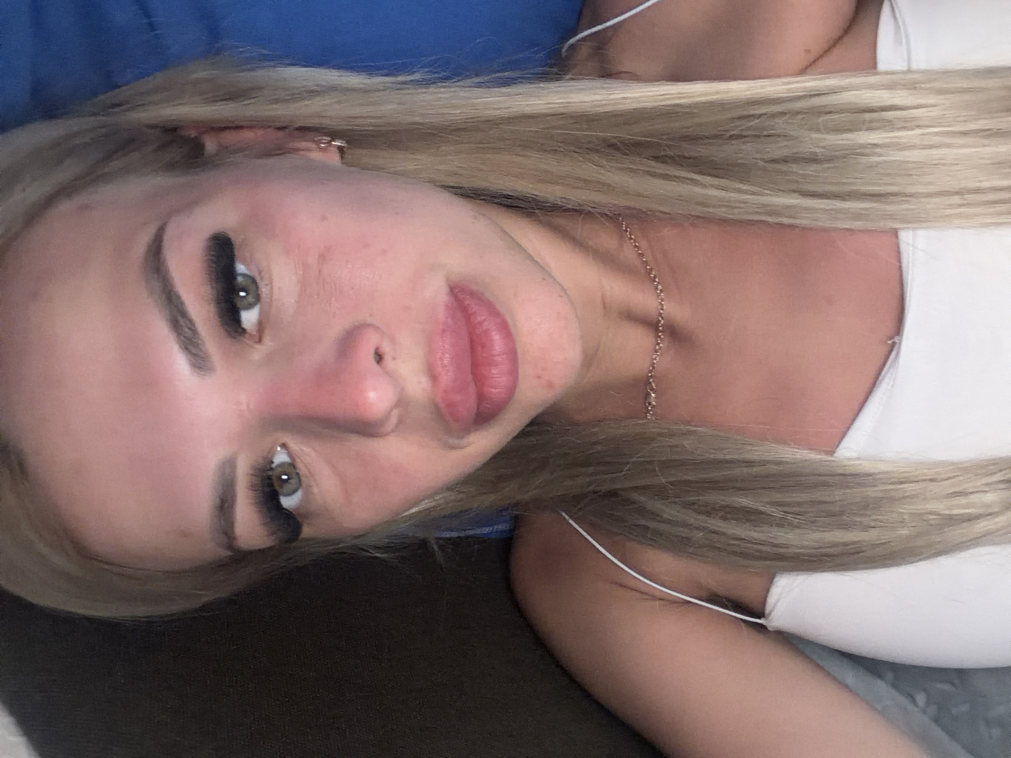 A close-up, selfie-style photograph features a blonde woman with fair skin, green eyes, and full lips. She is wearing a white tank top and a delicate gold necklace. The woman's face is turned slightly towards the camera, and her blonde hair is straight and falls past her shoulders. Her makeup includes false eyelashes and lip gloss. The background is blurred, but appears to be indoors with a dark area on the left and a blue surface on the right. The lighting is warm, highlighting her skin and creating a slight redness on her cheeks. There is no specific identifiable location within Kyiv, Ukraine, visible in the image.