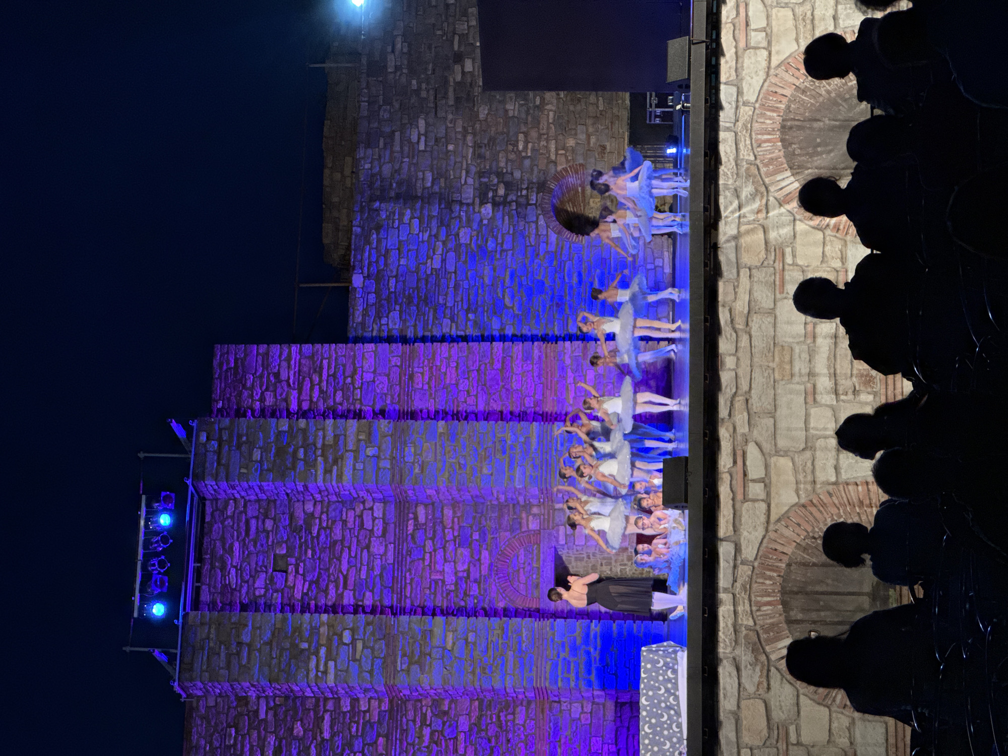 The image depicts an outdoor evening performance at the Tsarevets Fortress in Veliko Tarnovo, Bulgaria. On a raised stage, a group of young ballerinas in white tutus are performing, led by an adult instructor in a black dress. The backdrop is a stone wall illuminated with vibrant purple and blue lights. In the foreground, silhouetted figures of an audience are seated in chairs, observing the performance. The overall scene suggests a cultural event taking place within the historic fortress.