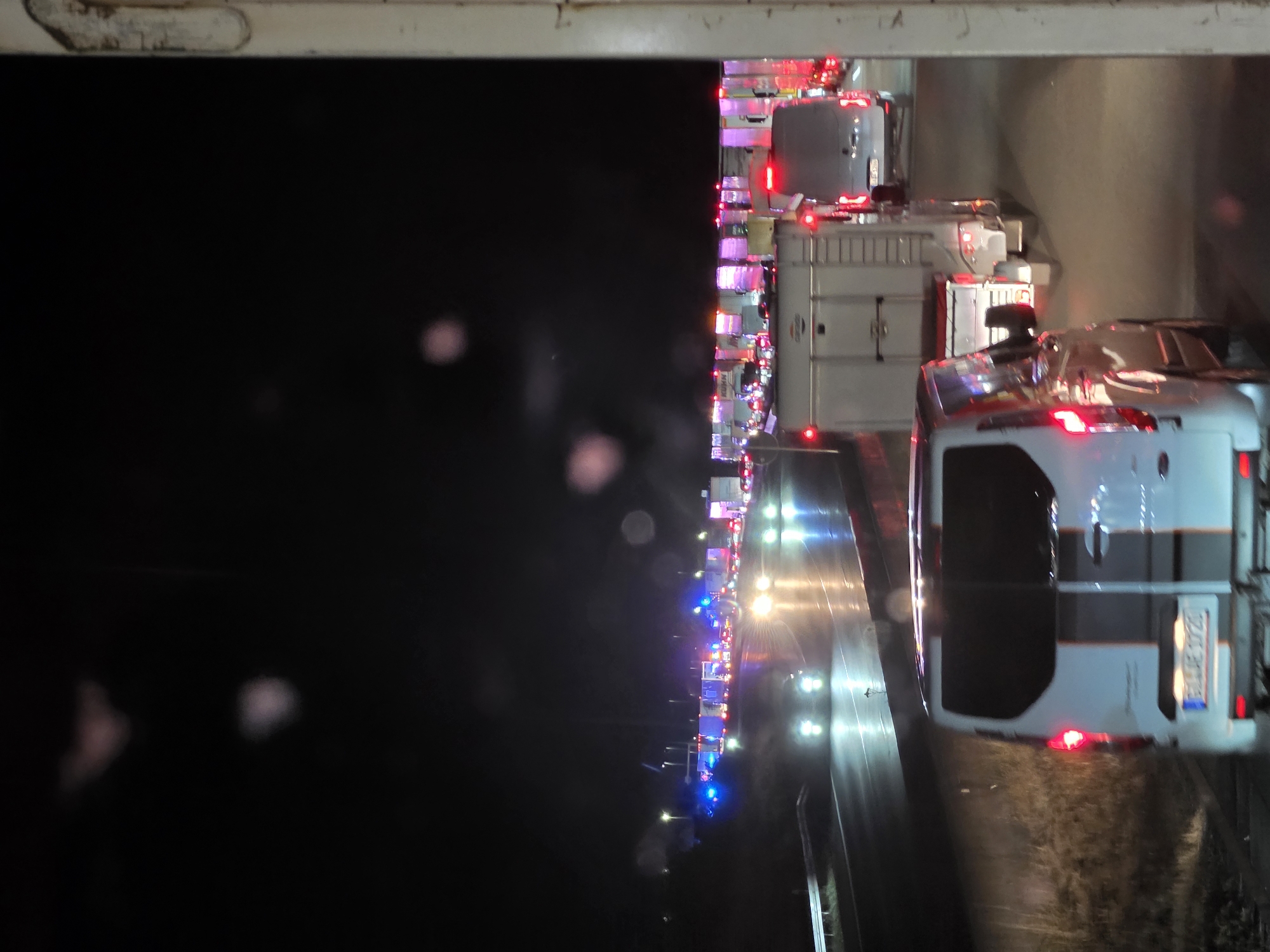 The image depicts a nighttime traffic scene on a highway, likely near Uettingen, Germany. A line of vehicles, including vans and trailers, are stationary or moving slowly, indicating a traffic jam. The road surface appears wet, reflecting the headlights of the vehicles and the lights from a distant structure that may be a toll booth or service area. Several vehicles are illuminated with flashing blue and red lights, suggesting the presence of emergency services. The sky is dark, with a few blurry lights visible, possibly from streetlights or distant buildings. The foreground shows the rear of a white van. The overall atmosphere is one of disruption and potential incident due to the emergency lights and traffic congestion.