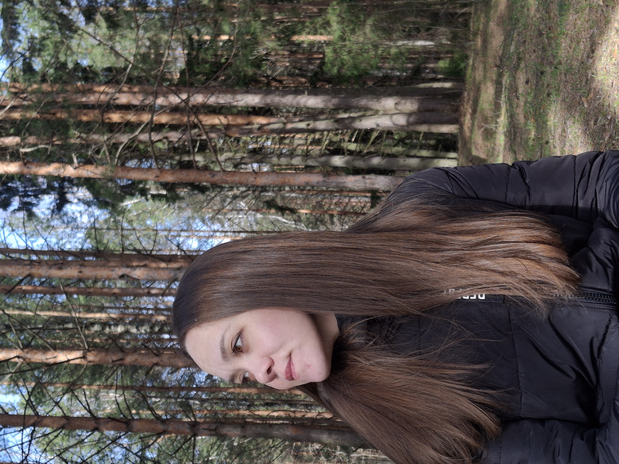 A portrait-style image features a young individual, likely female, with long, straight, dark brown hair, set against the backdrop of a dense pine forest. The person is wearing a black puffer jacket and is positioned slightly off-center to the right, looking downwards and towards the right with a soft, contemplative expression and a subtle hint of a smile. Her brown eyes are visible.
The background consists of numerous tall, slender pine tree trunks, indicating a forest environment. Patches of green foliage from the pine needles are visible through the branches, suggesting a vibrant, natural setting. The lighting appears to be natural daylight, possibly late morning or early afternoon, casting soft illumination without harsh shadows. The sky, visible in glimpses between the treetops, suggests clear weather. The overall ambiance is peaceful and serene.
On the black puffer jacket, a partial word in white text is visible, appearing to be "DERE" followed by obscured letters, possibly on a label or internal lining. The location is contextualized as Chaikovsky, Russia.