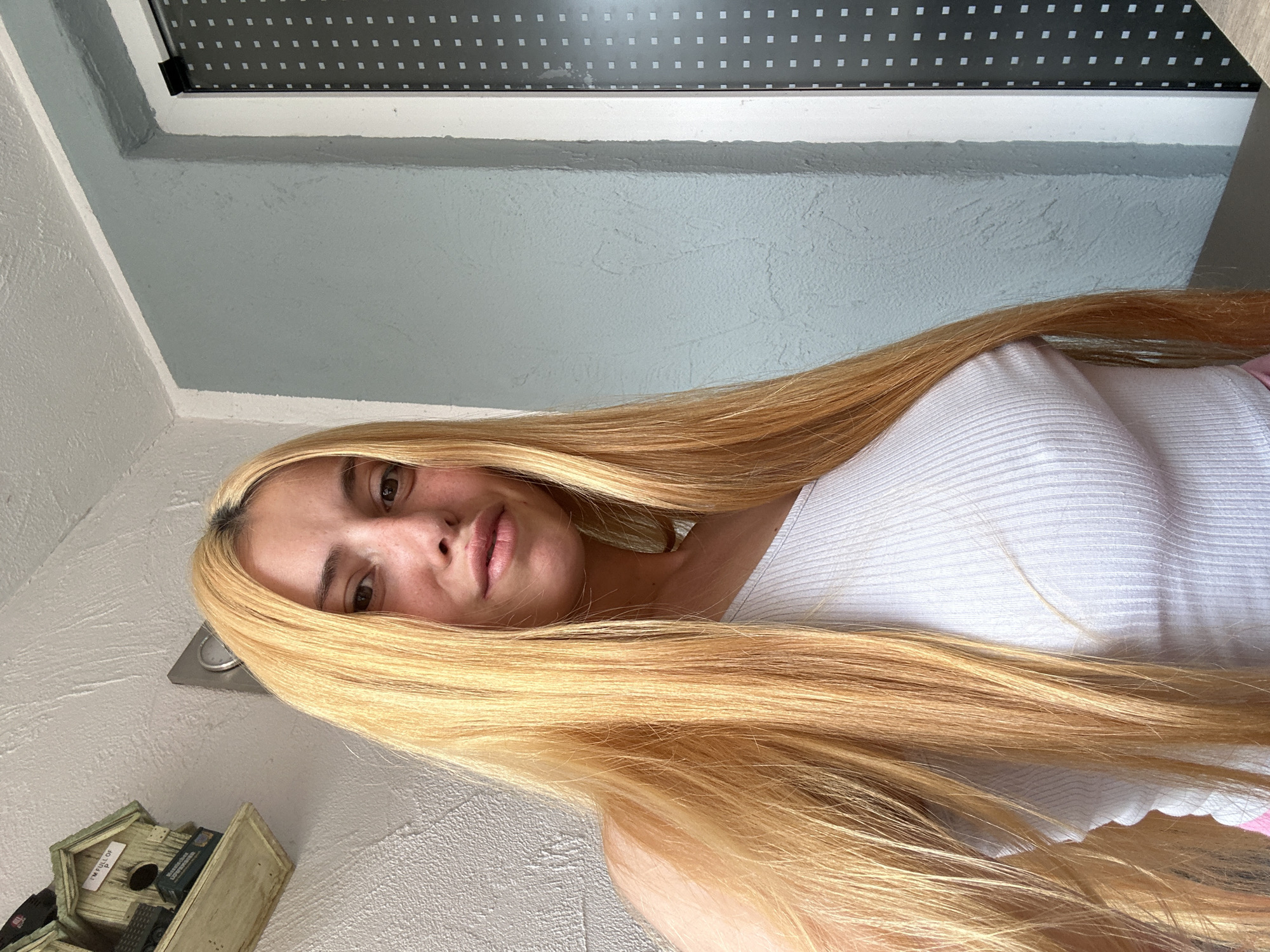 The image presents a blonde woman wearing a white ribbed tank top. She is indoors, with a window visible in the background. The wall color is a light grey. In the lower left corner, there is a small, decorative birdhouse. The location is specified as Heinebach, Germany, though no details in the image confirm this.