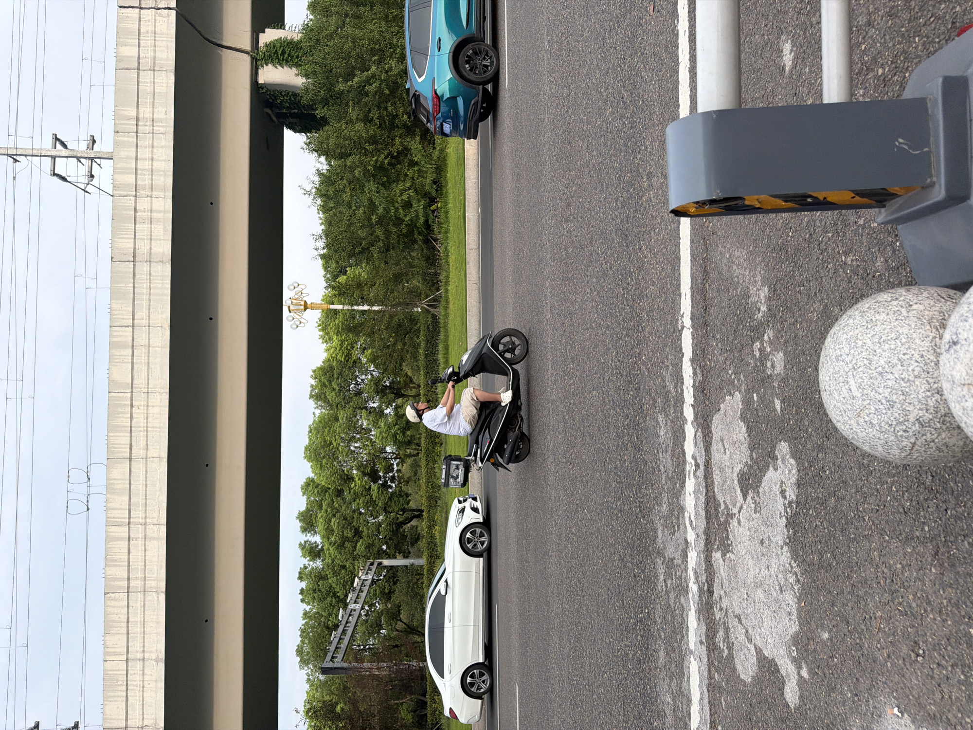 In the city of Jiangjin, China, a man wearing a white helmet and a white t-shirt is riding a black electric scooter. He is wearing khaki shorts and white shoes. The scooter has a black storage box attached to the rear. He is riding on a paved road with other vehicles present. To his left, a white Mercedes-Benz sedan is driving in the same direction. To his right, a teal SUV is also driving in the same direction. An elevated concrete structure is visible in the background, possibly an overpass or bridge, with overhead power lines. Lush green trees and foliage line the side of the road. The sky is overcast. In the foreground, a portion of a road barrier with yellow and black reflective tape is visible, along with some round granite decorative objects. The scene appears to be on an urban road during the daytime.