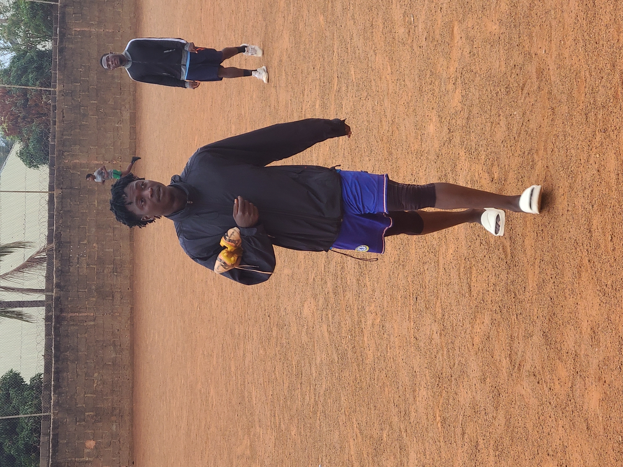 A man is standing on a red clay surface, possibly a tennis court, facing the camera. He wears a black jacket, blue shorts, and white sandals. He holds something yellow in his hand. In the background, another man is visible wearing a blue jacket and shorts. The location appears to be outdoors, with a brick wall on one side and trees visible in the background. The scene is in Barakin Lerawi, Nigeria.