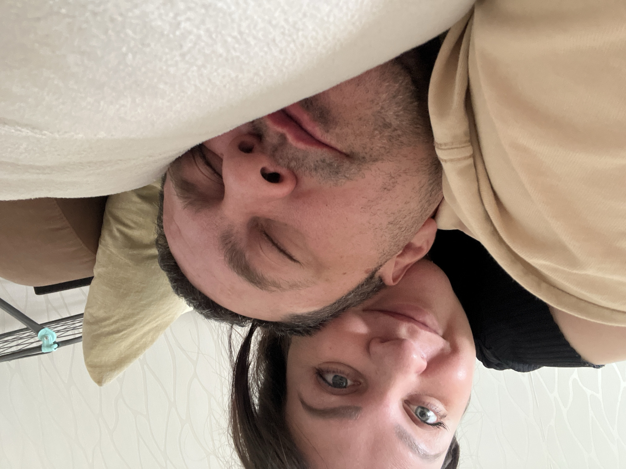 The image shows a man and a woman lying down together. The man appears to be asleep, with his eyes closed and face partially covered by a white blanket. The woman is awake and smiling, her face visible and turned towards the camera. The man has short, dark hair and stubble. The woman has dark hair pulled back and fair skin. They are lying on a beige pillow. The background shows a white wall with a subtle, patterned design and a metal rack with blue attachments. There is no evidence to confirm this image was taken in Perm, Russia.