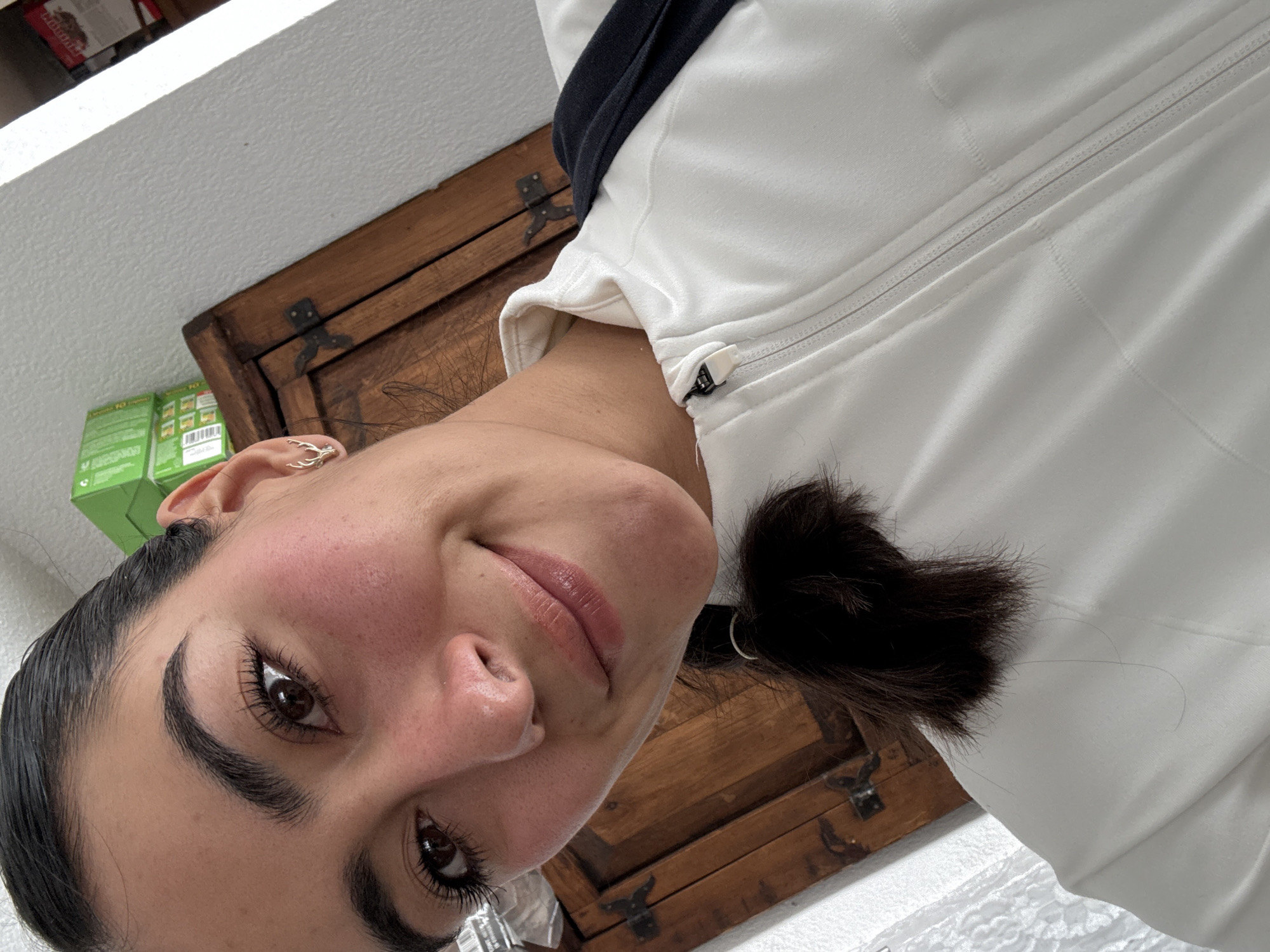 A close-up, tilted selfie of a woman with dark hair pulled back in a short ponytail. She has dark eyebrows and eyelashes, and her skin appears flushed. She is wearing a white zippered jacket with a black collar. Behind her is a wooden chest with metal hinges and part of a white wall or surface. Part of a green product box is visible in the upper left corner. The location is indeterminate from visual information, and cannot be ascertained to be Puebla, Mexico.