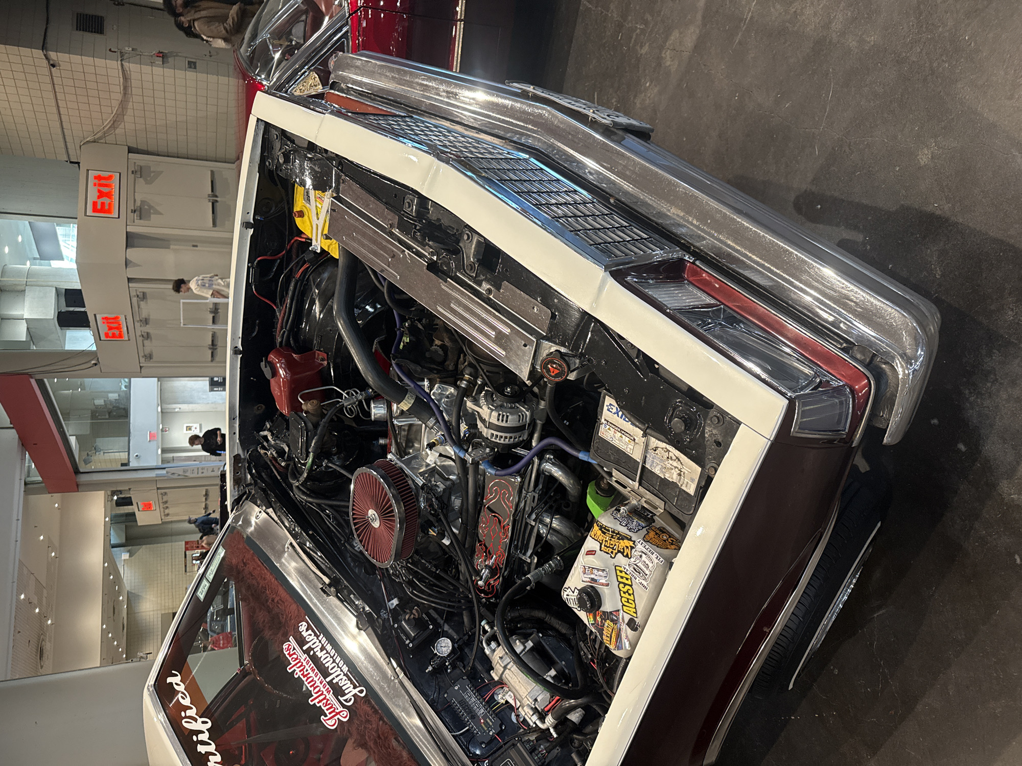 A high-angle, tilted, indoor shot shows the open hood of a burgundy and white vintage car, revealing the engine. The car is parked on a smooth, dark gray floor. The engine compartment is densely packed with various components, including a red air filter, a battery covered in stickers, and numerous wires and hoses. The hood and front end of the car are visible, featuring chrome details and a white stripe.
In the background, a portion of a building is visible, suggesting a large indoor space, possibly a convention center or a garage. An exit sign is visible on the wall. The location is presumed to be New York City based on the prompt. The overall scene suggests a car show or an exhibition of vintage and customized vehicles.
