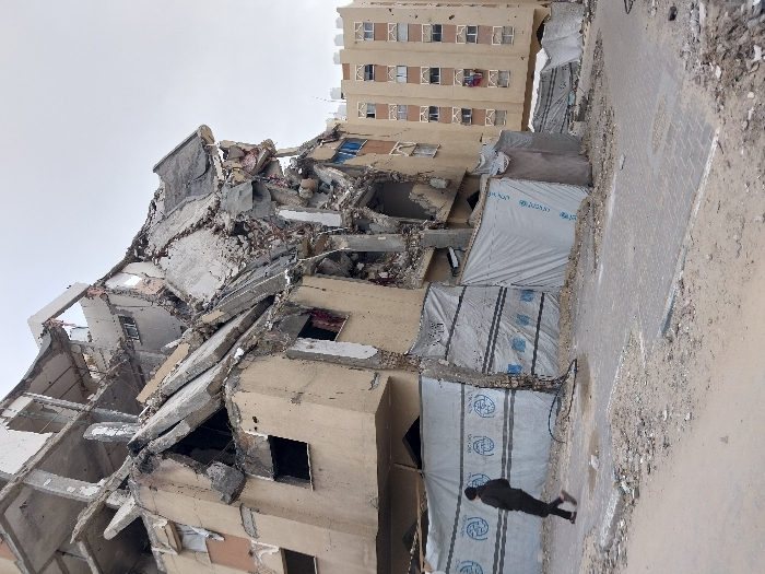 An outdoor scene in Khan Yunis, Palestinian Territory, depicts a significantly damaged urban environment under an overcast sky. In the foreground, a paved sidewalk and dirt ground covered in rubble lead towards a multi-story building on the left that has suffered a catastrophic partial collapse. Its concrete structure is severely compromised, with shattered slabs, exposed rebar, and twisted elements cascading downwards and inwards, indicating extensive destruction, particularly in its upper and central sections. The lower levels of this building are partially obscured by temporary barriers constructed from light grey tarpaulin material, some bearing blue circular logos. A lone individual, dressed in dark clothing, walks from left to right across the paved sidewalk in front of these barriers, their head slightly bowed. To the right of the collapsed structure, in the midground and background, stands another multi-story building with a light beige facade and numerous windows, showing visible pockmarks and minor surface damage. Debris is scattered across the ground in the immediate foreground and on the sidewalk.