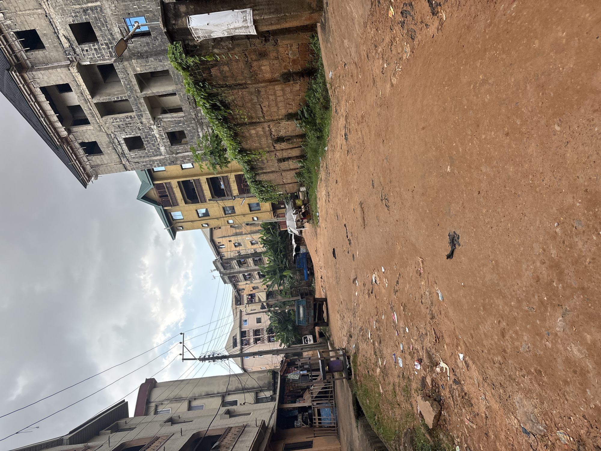 The image depicts a dusty, unpaved street in the city of Onitsha, Nigeria, likely in a densely populated residential area. Multiple multi-story buildings, some appearing to be under construction or in a state of disrepair, line the street. The buildings are varied in color and architectural style, with some featuring balconies and exposed concrete. A prominent feature is a tall, uncompleted building on the right side of the street, constructed with cinder blocks and lacking windows. A crumbling brick wall, partially covered in vines, separates the street from this building.
The street itself is covered in reddish-brown dirt and littered with various debris, including plastic bags, paper, and other refuse, indicating a lack of proper waste management. Several vehicles are visible: a small blue truck, a dark green flatbed truck with a trailer, and what appears to be a white van parked further down the street. A makeshift stall or shop with wooden structures and a display of goods is situated on the left side of the street. Overhead, numerous electrical wires crisscross the sky, supported by poles, a common sight in many Nigerian cities. The sky is overcast with grey clouds, suggesting an impending rain or a generally cloudy day. No people are clearly visible in the image, though the presence of a shop and vehicles suggests human activity in the area. The overall impression is one of a busy, somewhat neglected urban environment.