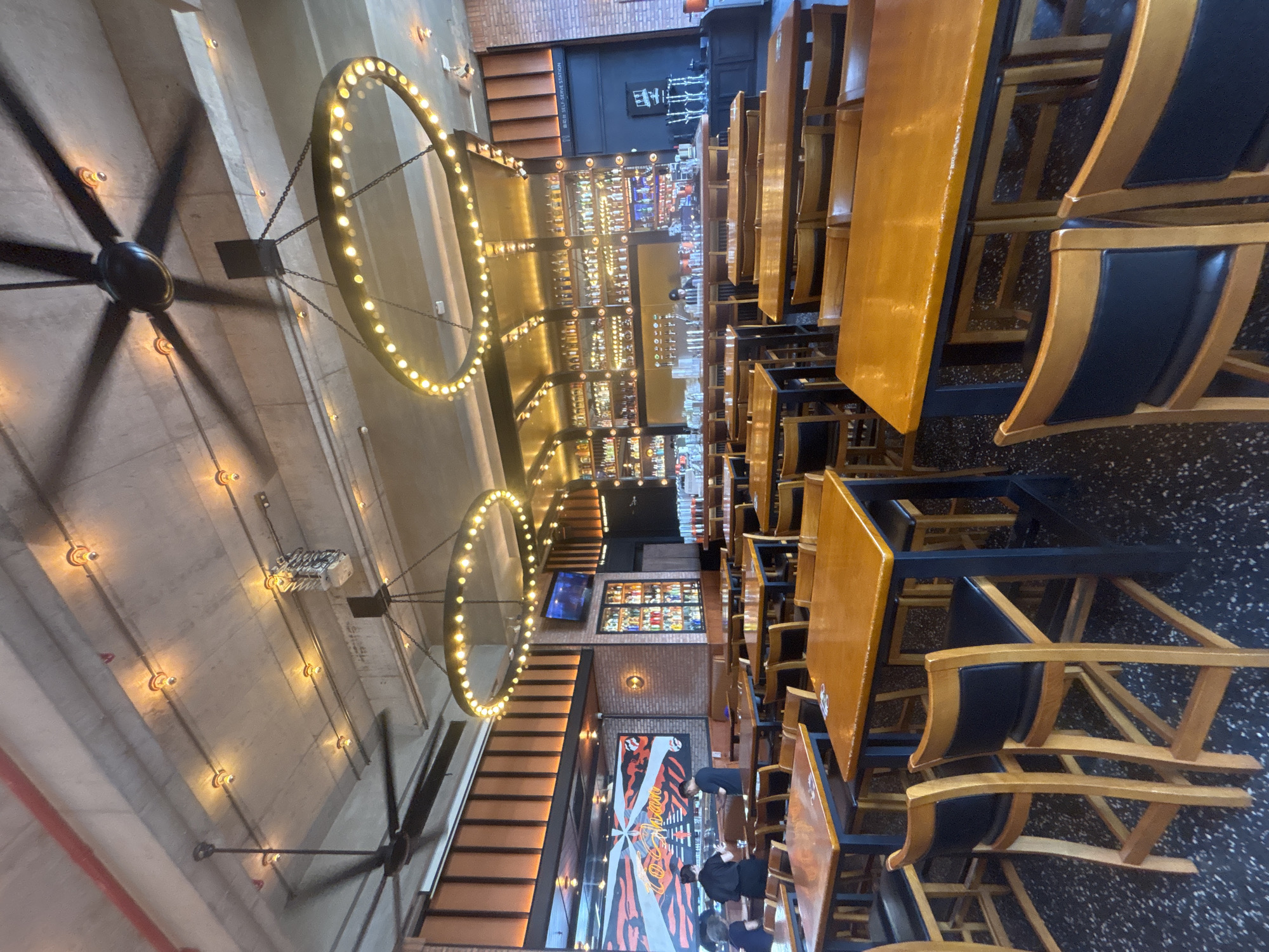 The image captures the interior of a restaurant in Nanhai District, China, during daylight hours. The restaurant features an industrial-chic aesthetic with exposed concrete ceilings, ductwork, and brick walls. Large circular chandeliers adorned with numerous bulbs hang prominently, complementing the industrial ceiling lights. The space is furnished with numerous wooden tables and chairs, including some high-top tables with bar stools. A well-stocked bar area is visible in the background, featuring rows of bottles and a bartender attending to customers. The atmosphere appears casual and inviting, suggesting it is either early evening or the restaurant is preparing for its dinner service.