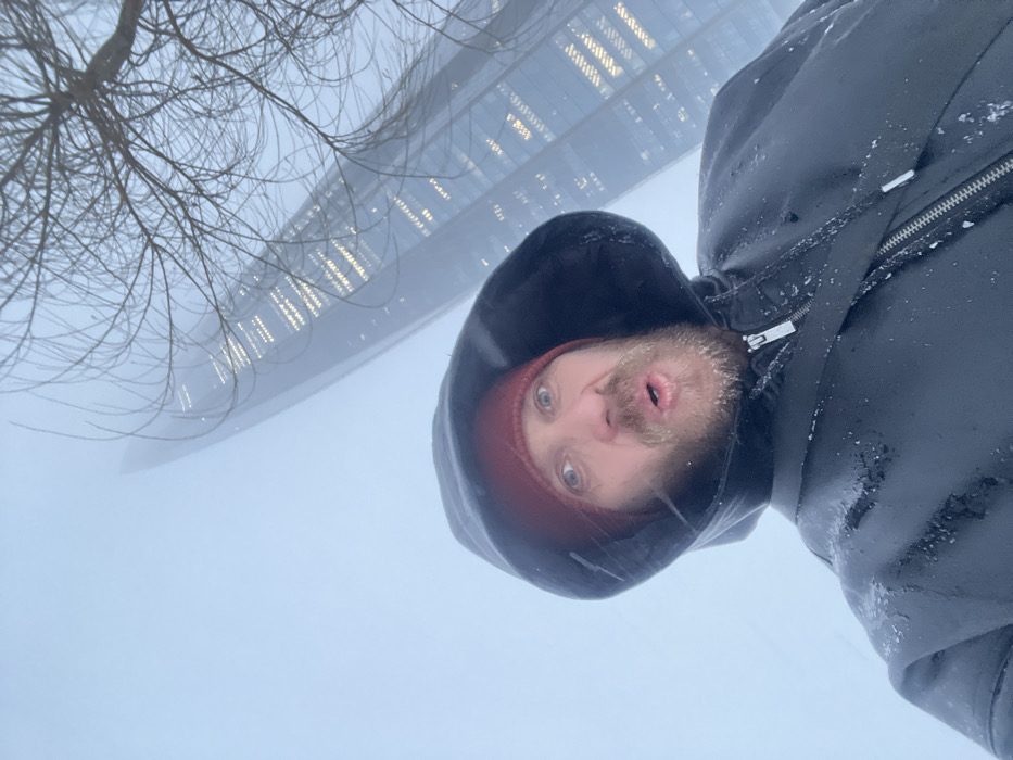A male subject is depicted in a close-up, selfie-style photograph taken in Saint Petersburg, Russia, with the Lakhta Center skyscraper prominent in the background. The man, positioned centrally in the foreground, wears a dark hooded jacket and a dark red beanie. His beard and the surface of his jacket show visible accumulation of snow. He looks directly at the camera with wide eyes and his mouth slightly open. In the background, the Lakhta Center, a tall, tapering glass skyscraper with numerous illuminated windows, extends upwards and is partially obscured by dense fog or heavy snowfall. Bare tree branches are visible in the upper right portion of the frame. The overall environment indicates cold, snowy, and misty winter conditions.