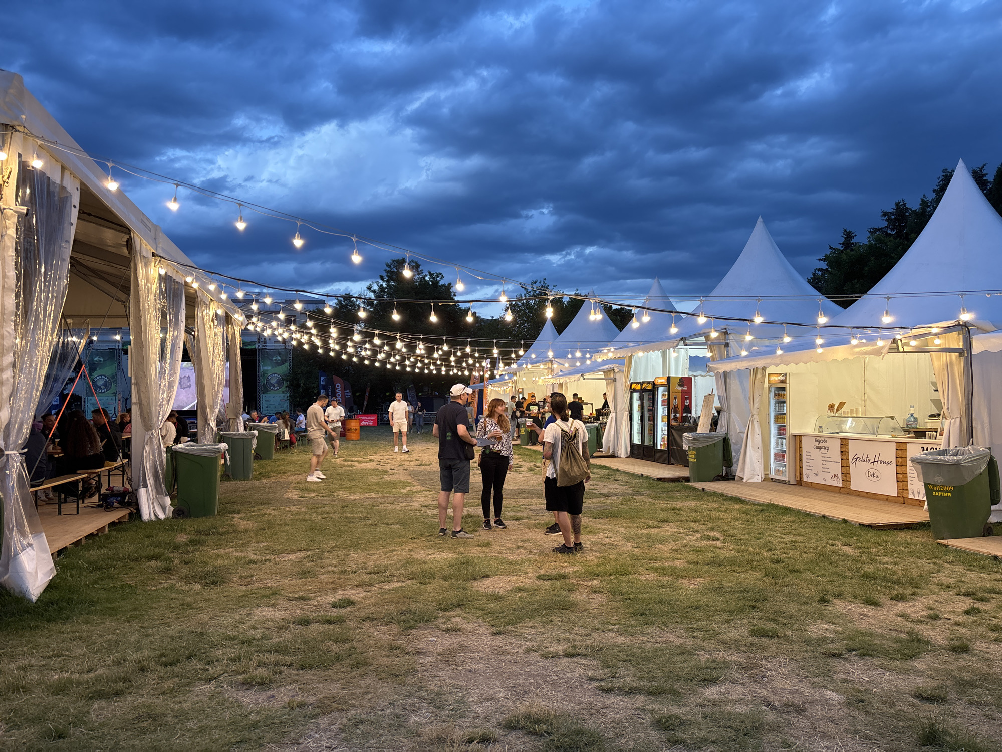 This is a photograph taken at dusk at an outdoor event, possibly a festival or market, in Sofia, Bulgaria. The sky is dark blue with heavy clouds, indicating approaching rain or a storm. String lights are illuminating the scene, creating a festive atmosphere. Several white tent structures are visible, some with transparent plastic sheeting acting as walls. People are milling about in the grassy area between the tents.
In the foreground on the left, a long wooden bench is set up under a tent, with several people sitting and standing. Next to the bench are two large green trash cans. Further into the scene, a group of four people are standing and conversing in the center of the image. One man wears a dark t-shirt and shorts, a white cap, and glasses. A woman stands next to him in a patterned shirt and dark pants. Another man is in a black t-shirt and shorts, carrying a backpack, and a fourth person, a man, is wearing a light-colored t-shirt and shorts.
To the right of the central group, there are more tents with open fronts, likely food or merchandise stalls. One tent clearly advertises "Gelato House" with an image of ice cream. Refrigerated display cases are visible inside. There are more trash cans scattered throughout the area. In the background, trees are visible beyond the tents, and further out, the lights of the city can be seen. The overall impression is of a lively event taking place in an outdoor setting as daylight fades.